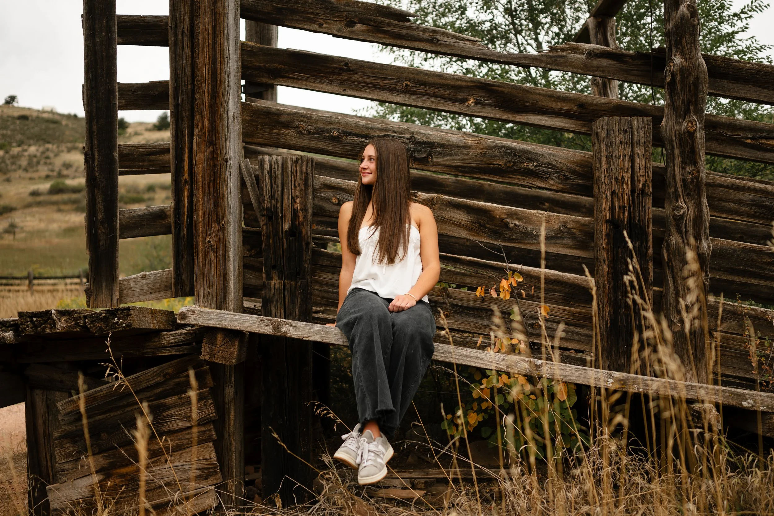 A high school senior sitting on a cattle chute in Fort Collins Colorado wearing western inspired jeans and a top looking over her shoulder during golden hour.