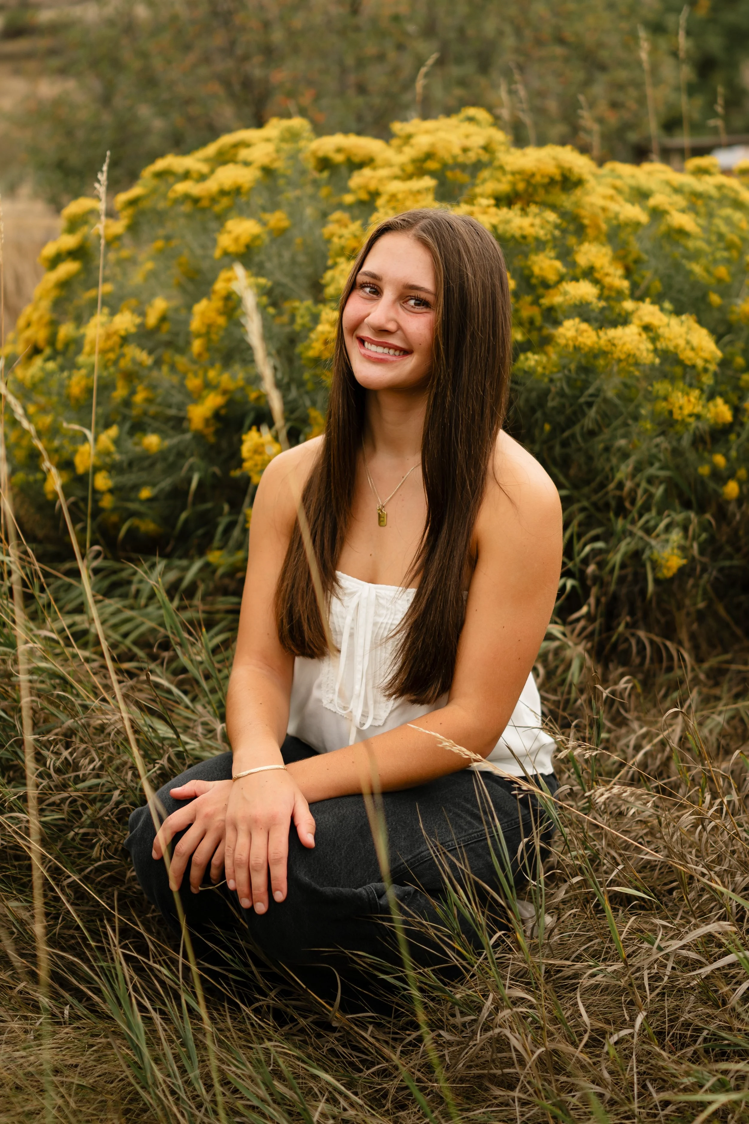 A high school senior girl wearing a strapless white top and black jeans is kneeling down in a field at Horsetooth in Fort Collins, Colorado. She is by yellow wild flowers with a western vibe during golden hour. 