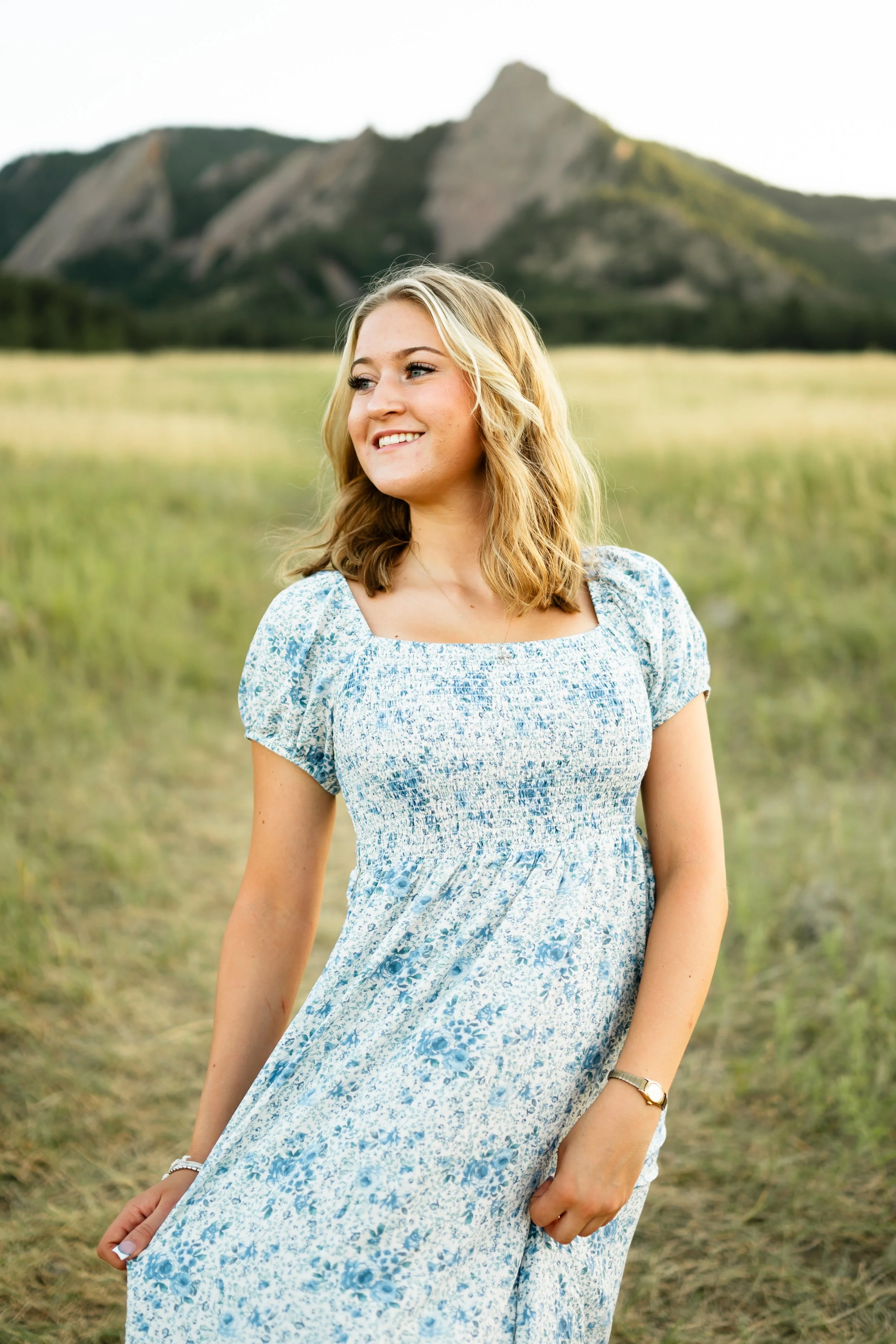 A high school senior girl at golden hour in Boulder Colorado at Chautauqua Park wearing a long blue dress with blonde hair looking over her shoulder while smiling.