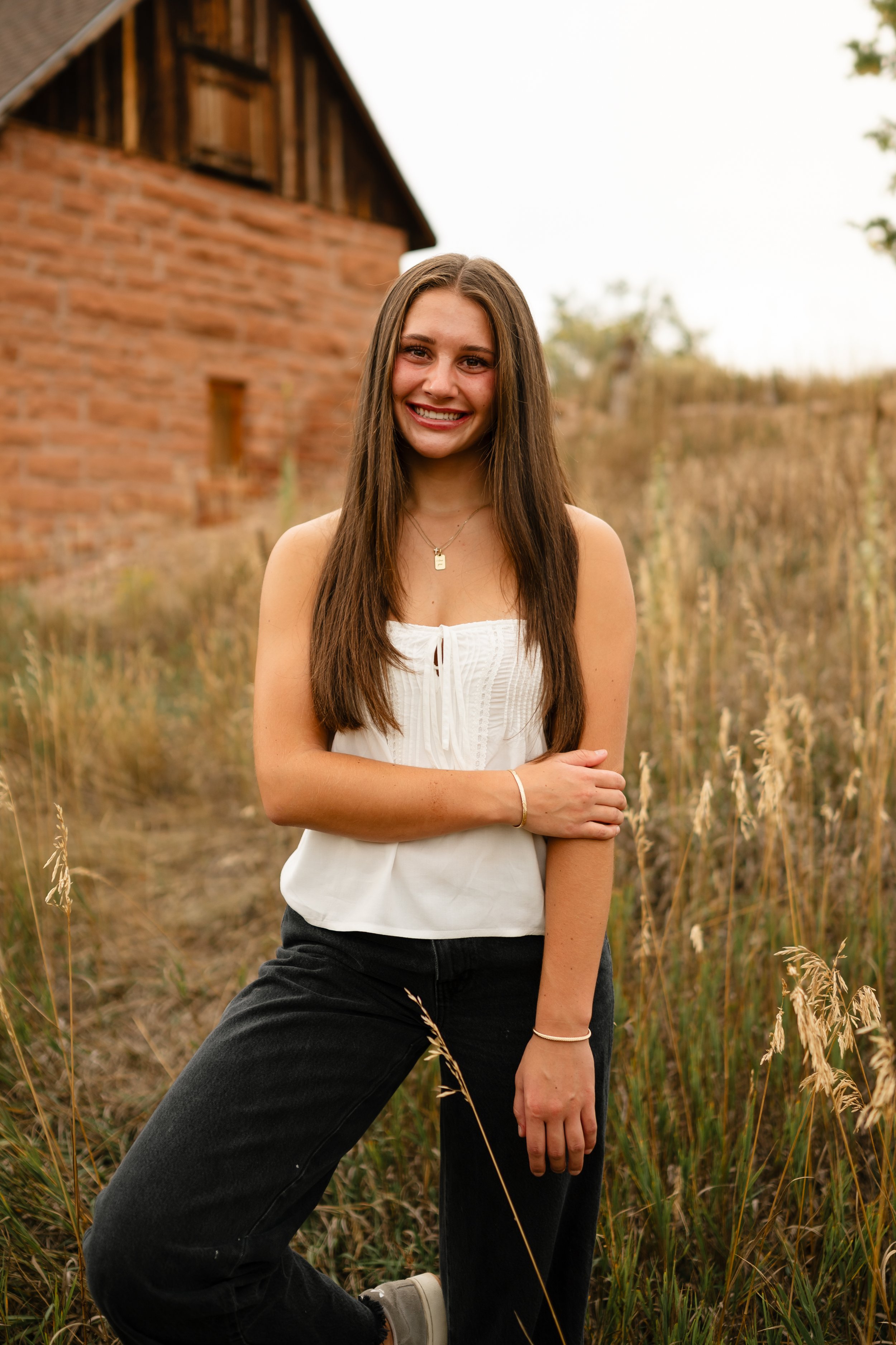 A high school senior is in a field at Horsetooth in Fort Collins, Colorado. She is wearing a white top and black jeans with her hands in her pockets and her legs crossed. She is standing in front of an old brick building with a western vibe. 