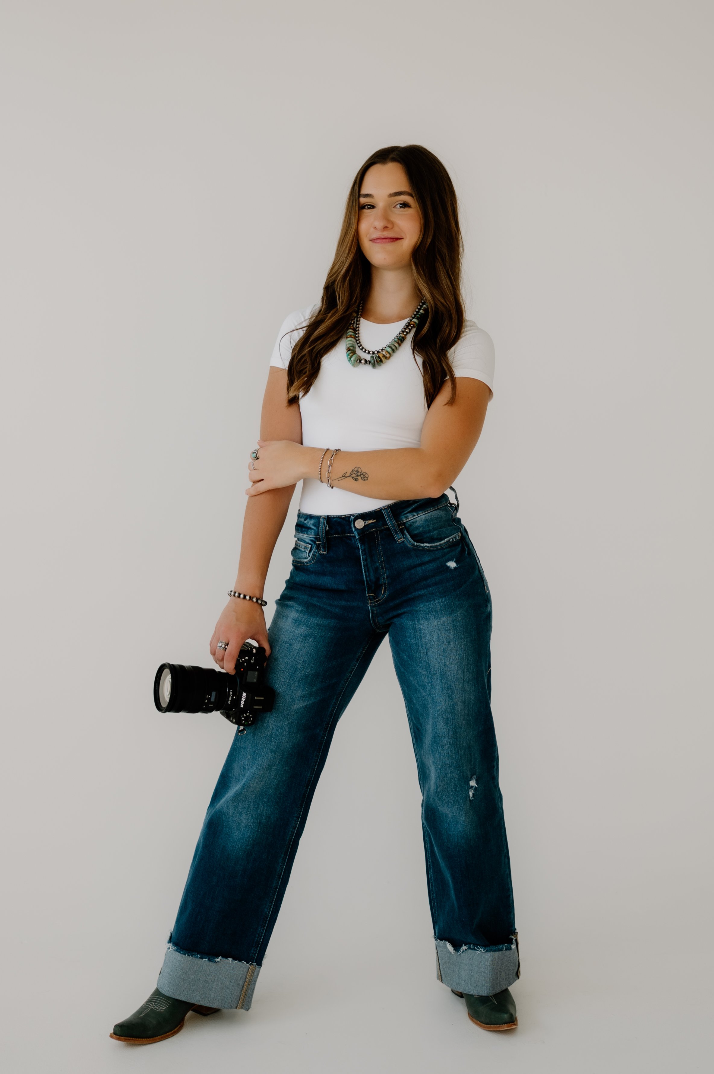A Northern Colorado photographer at Sixteenth Street Studio in Greeley Colorado holding a camera and dressed in western attire including jeans and cowboy boots with a white top and turquoise jewelry and Navajo pearls.