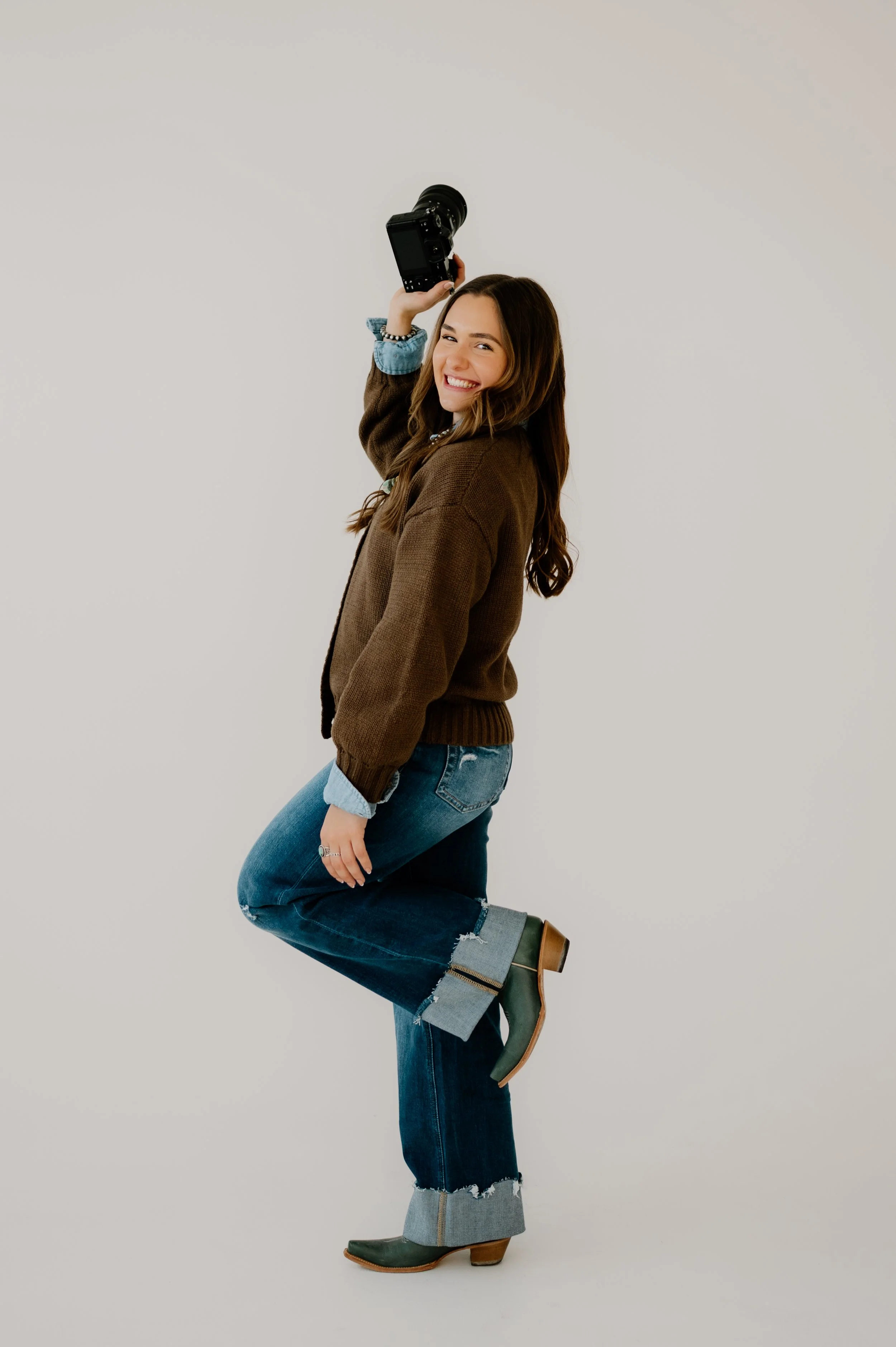 A Colorado wedding and portrait photographer at Sixteenth Street Studio in Greeley Colorado wearing jeans and cowboy boots, a brown sweater and denim button up holding a camera with Navajo pearls and turquoise jewelry on.