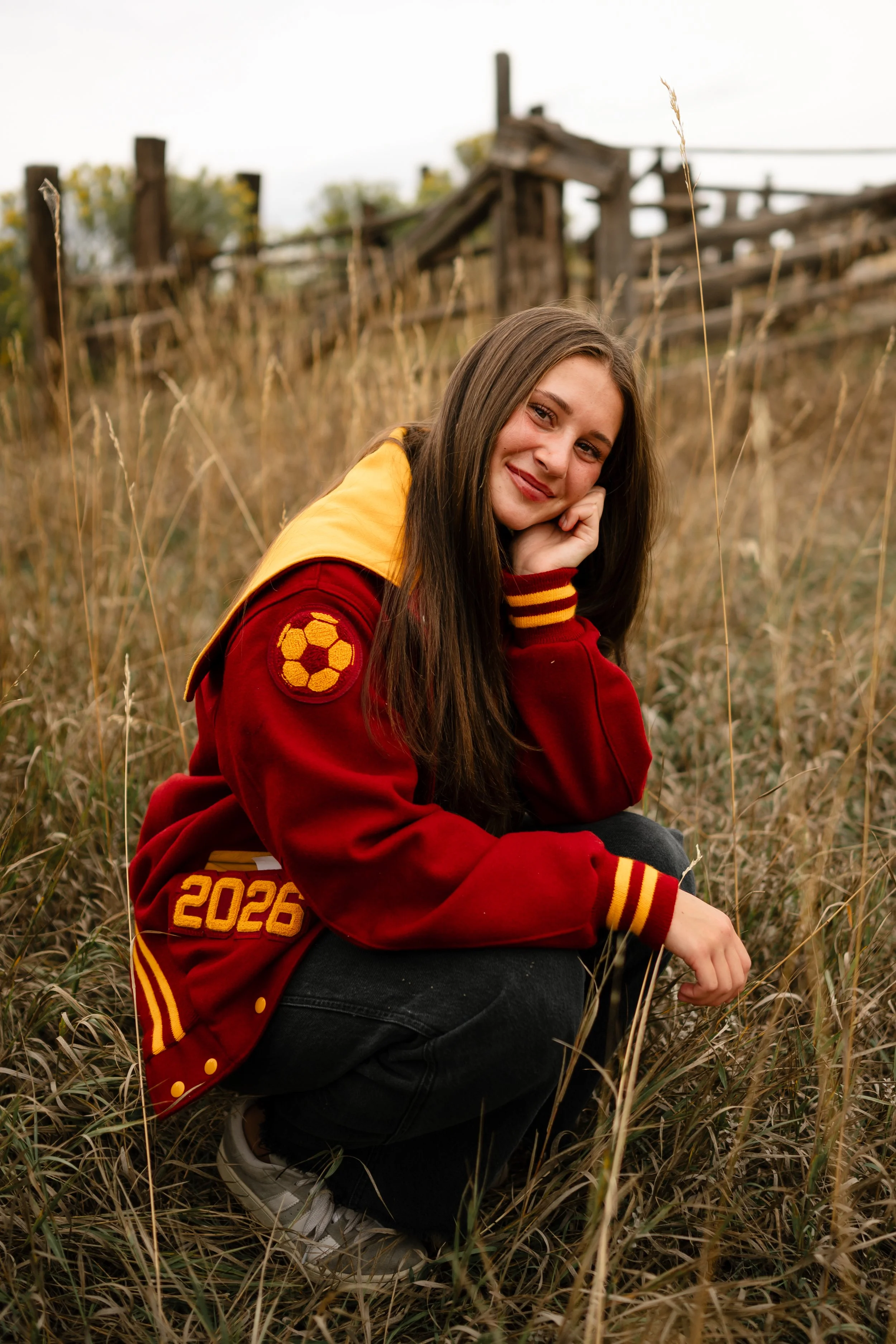 A high school senior girl wearing a Rocky Mountain High School letterman jacket with a soccer patch, squatting down in a field at Horsetooth in Fort Collins, Colorado with a western luscious green field behind her.