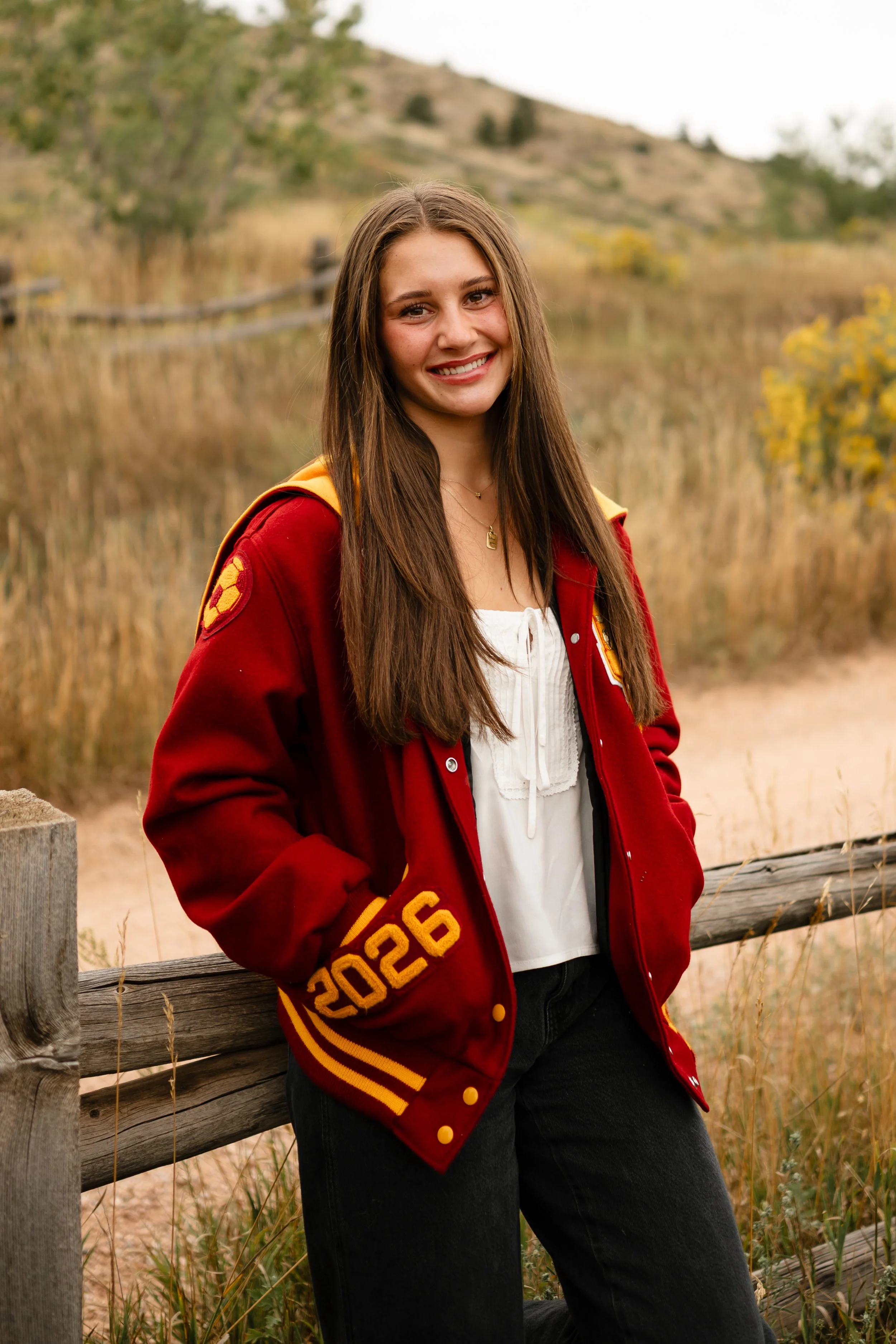 A high school senior girl wearing a Rocky Mountain High School letterman jacket leaning against a fence at Horsetooth in Fort Collins, Colorado. She is wearing black jeans and a white top during golden hour.