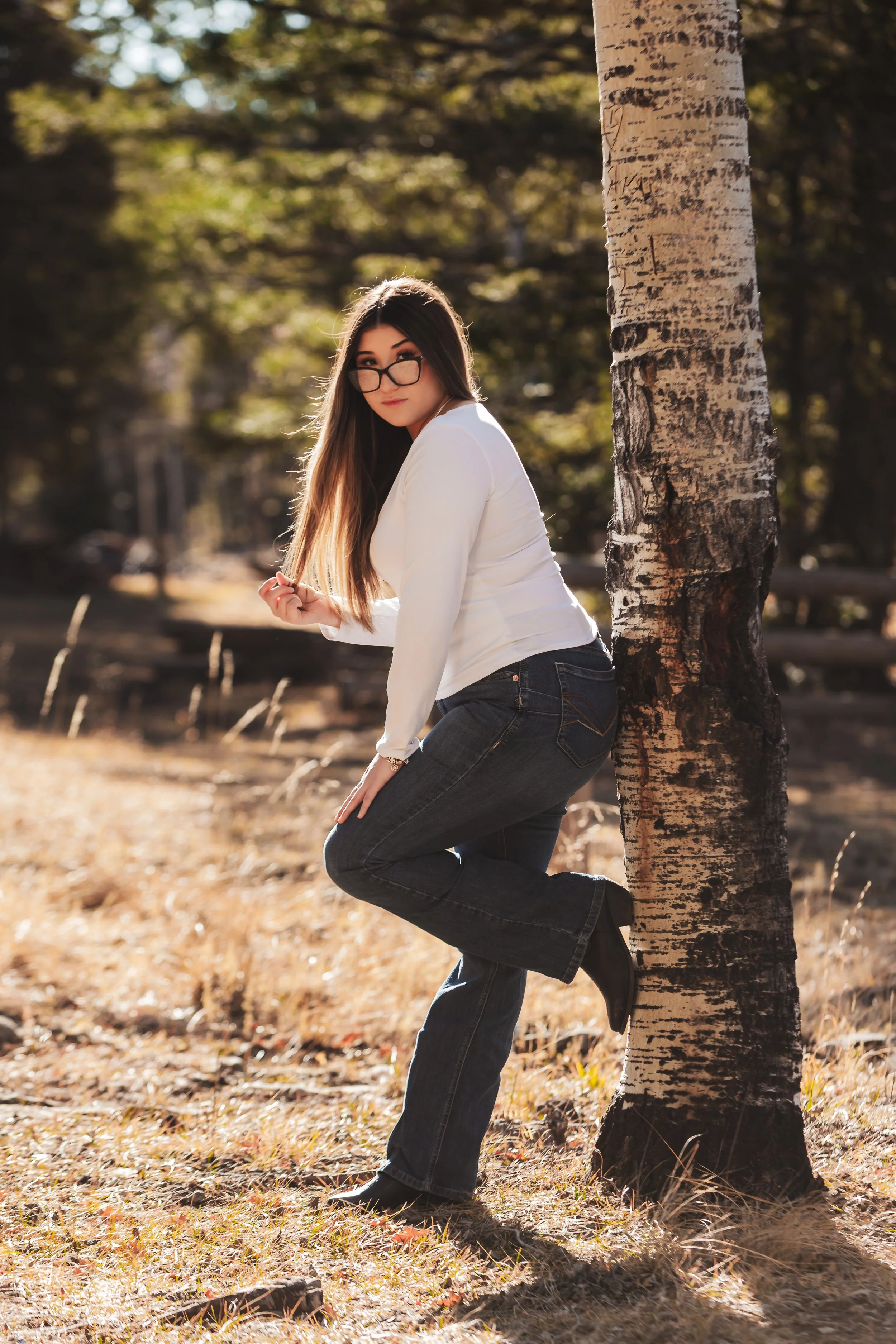 A young woman with long brown hair, wearing glasses, a white top, and dark jeans, leans against a tree in a forested area during autumn. Senior Rep photoshoot in Cloudcroft, NM.