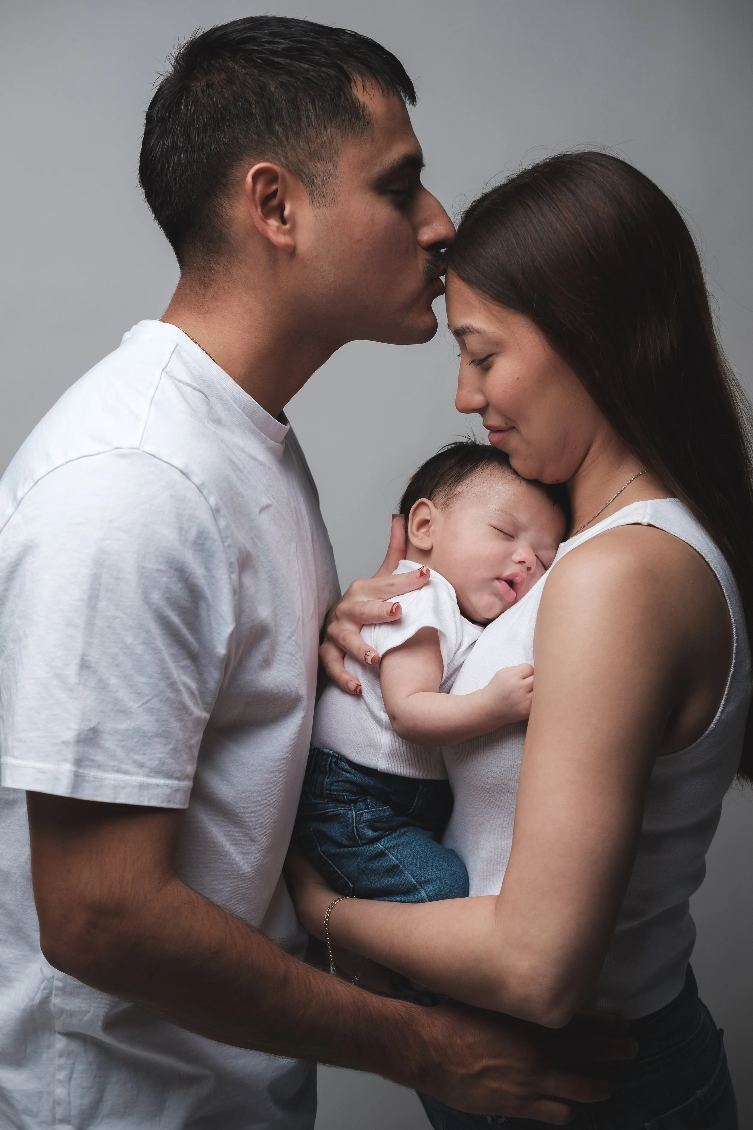 A man and a woman hold a sleeping baby close to each other, with the man kissing the woman's forehead. Home studio family photoshoot in El Paso, TX.
