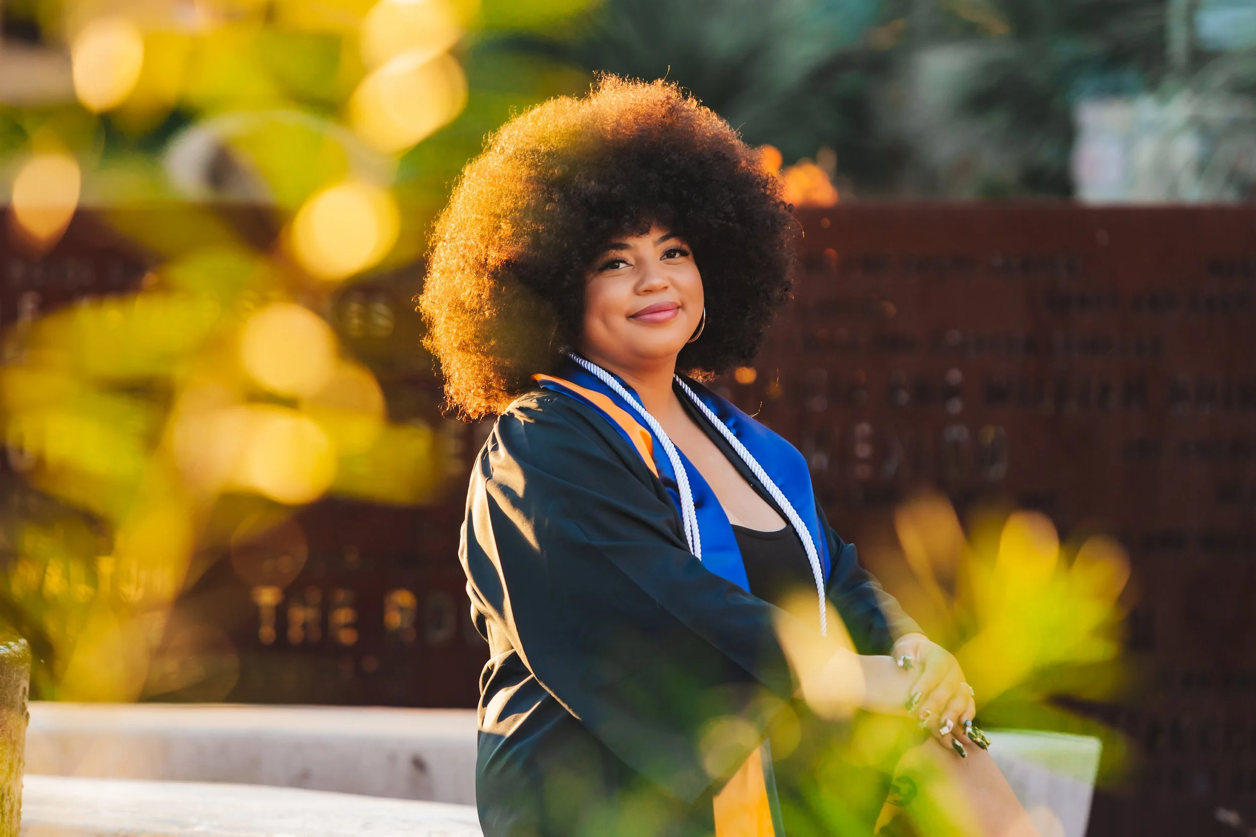 Young woman in a graduation gown and cords, smiling outdoors during sunset, with blurred green leaves in the foreground.