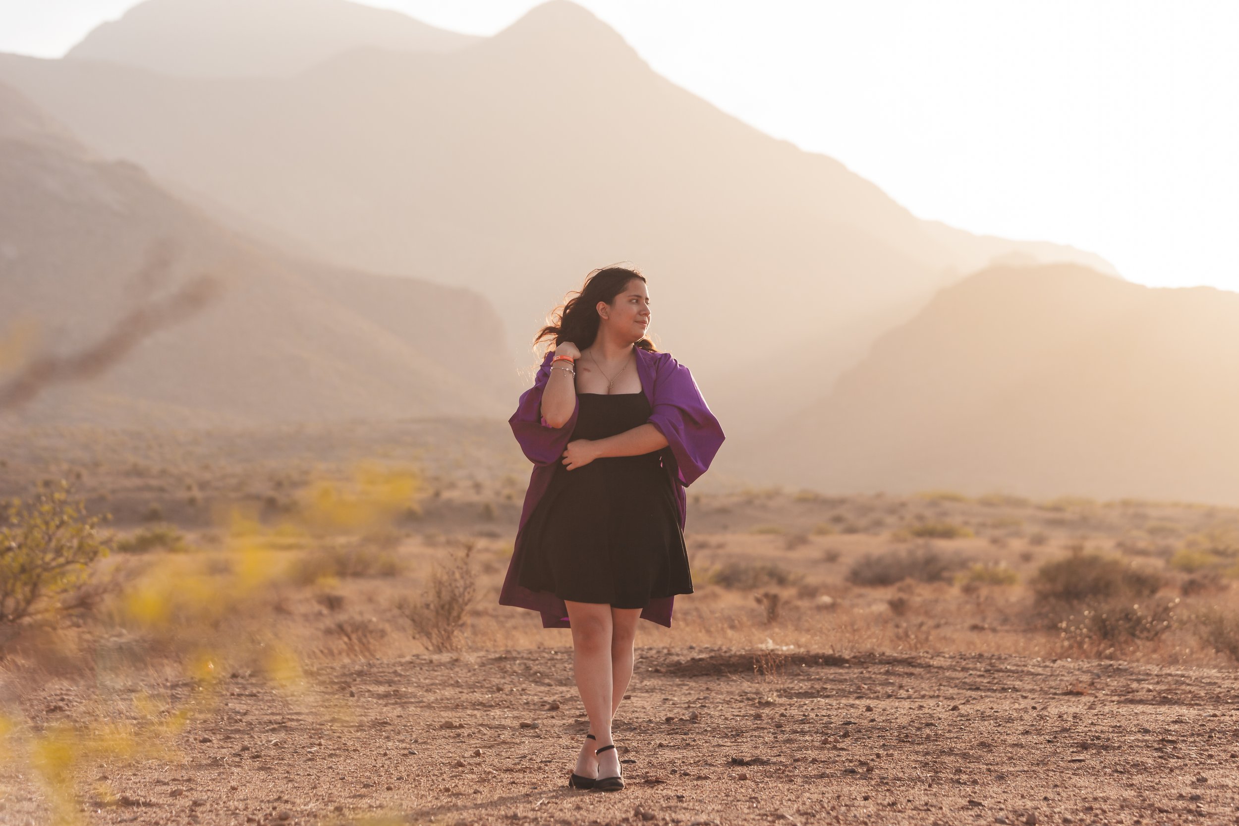 High School Senior girl in El Paso, TX near the Franklin Mountains at sunset wearing a black dress and purple graduation gown.