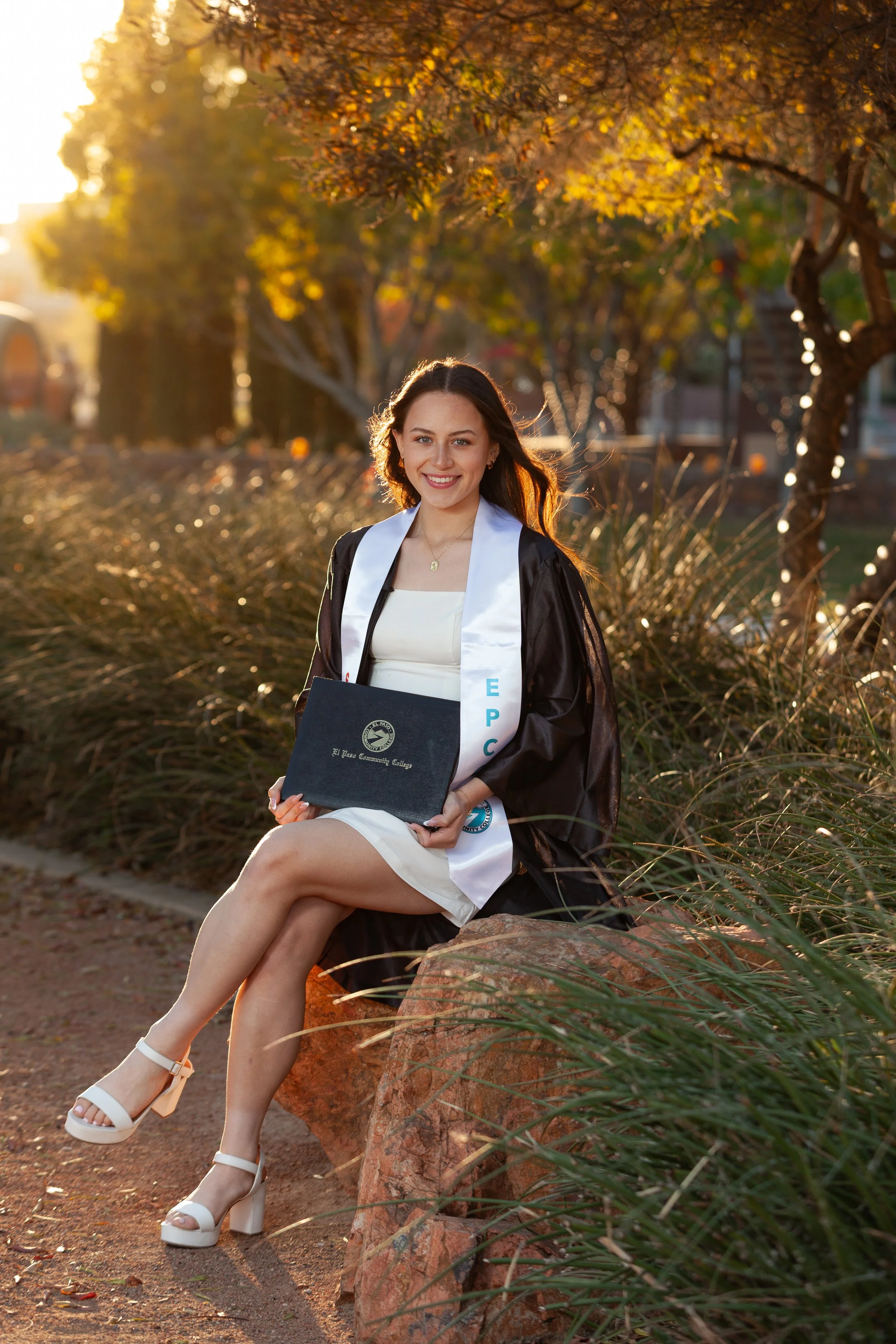 A young woman in a graduation gown and cap sitting on a rock outdoors during sunset, holding a diploma from El Paso Community College, smiling. Photoshoot at Texas Tech University Health Sciences Center at Sunset in El Paso, TX.