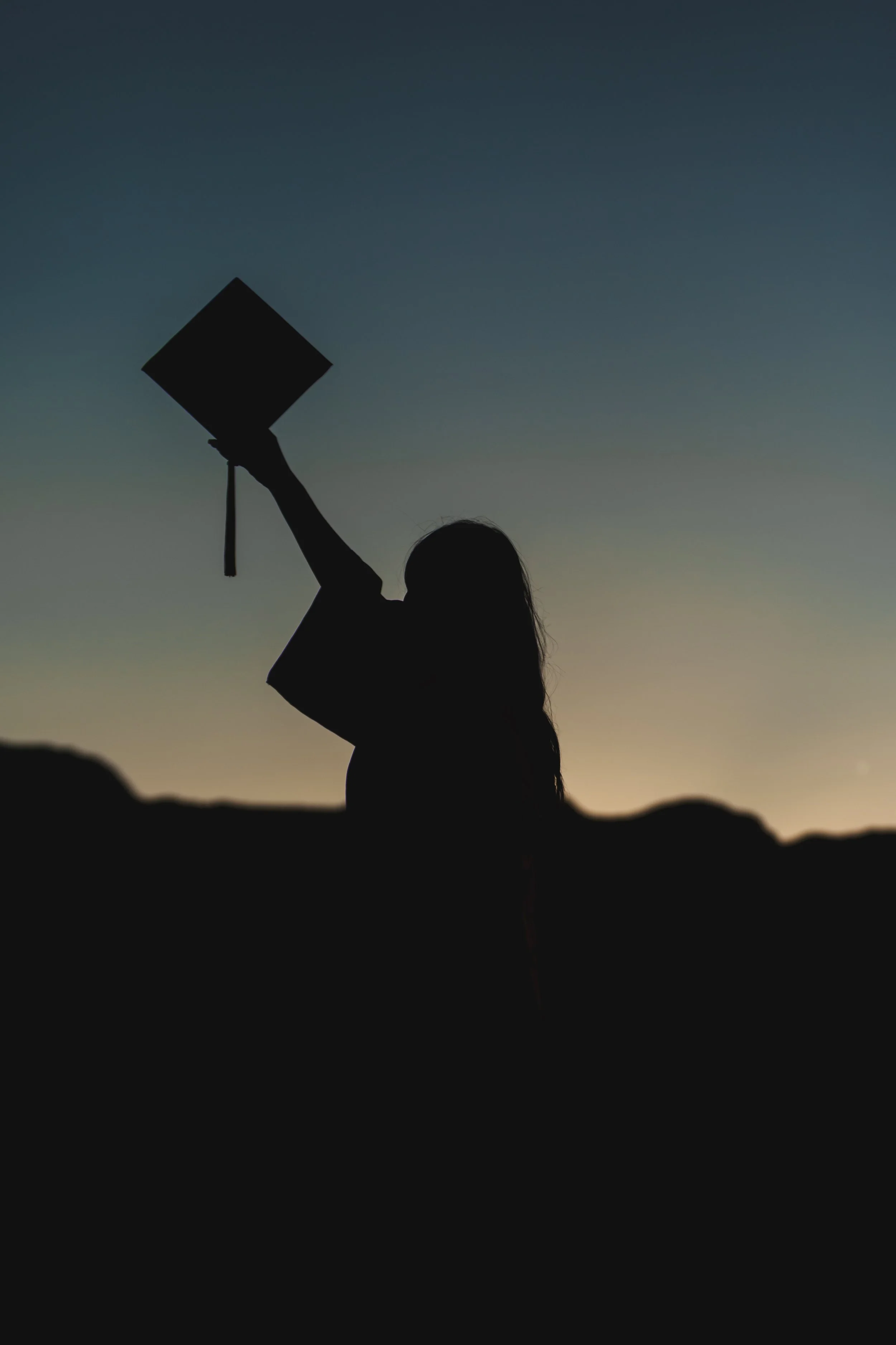 Silhouette of a person holding a graduation cap and diploma against a sunset sky. Outdoor senior photos in El Paso at sunset.