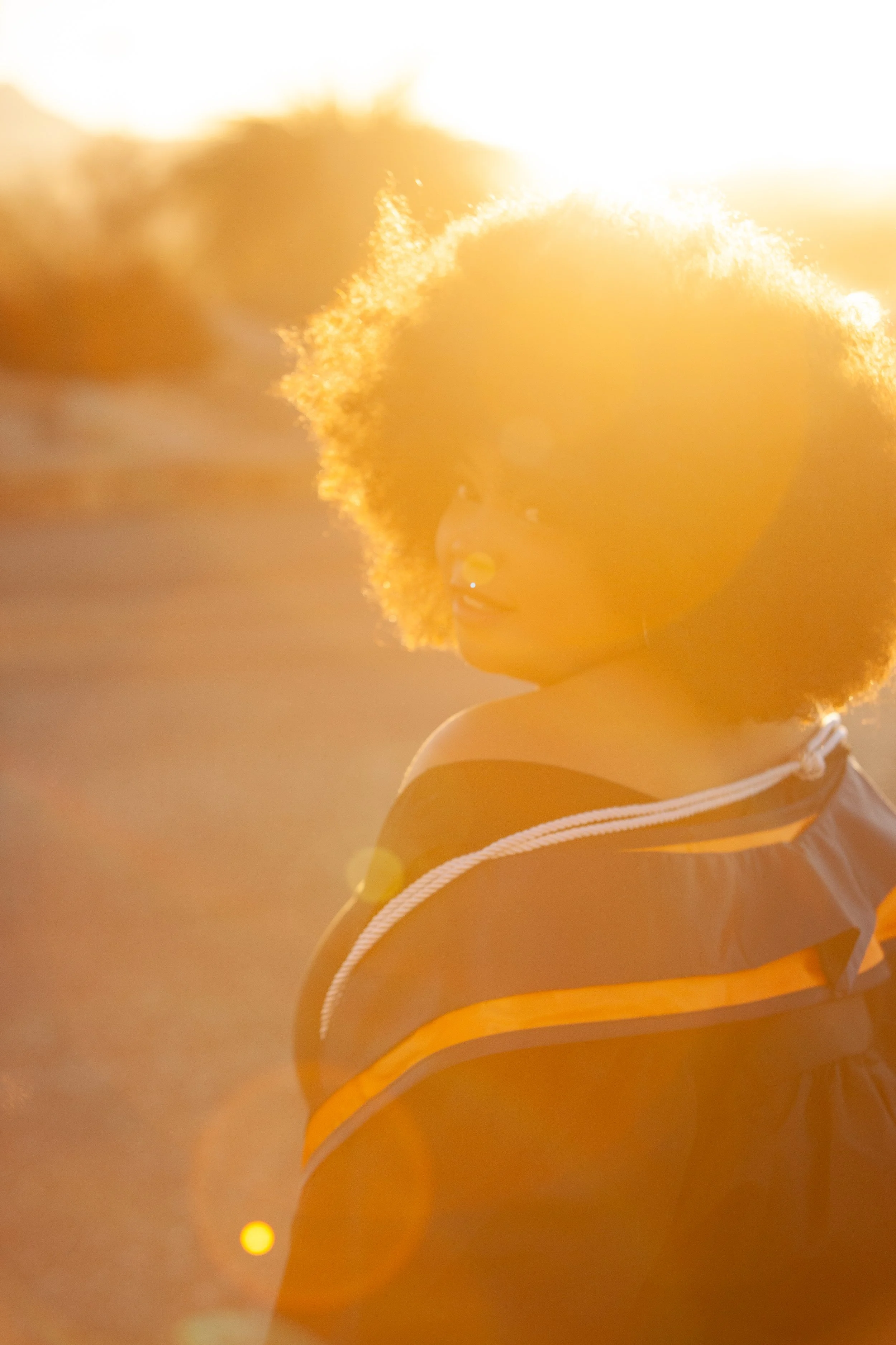 Young woman with a large afro hairstyle smiling with sunlight backlighting her, wearing off-shoulder clothing, outdoors during sunset.