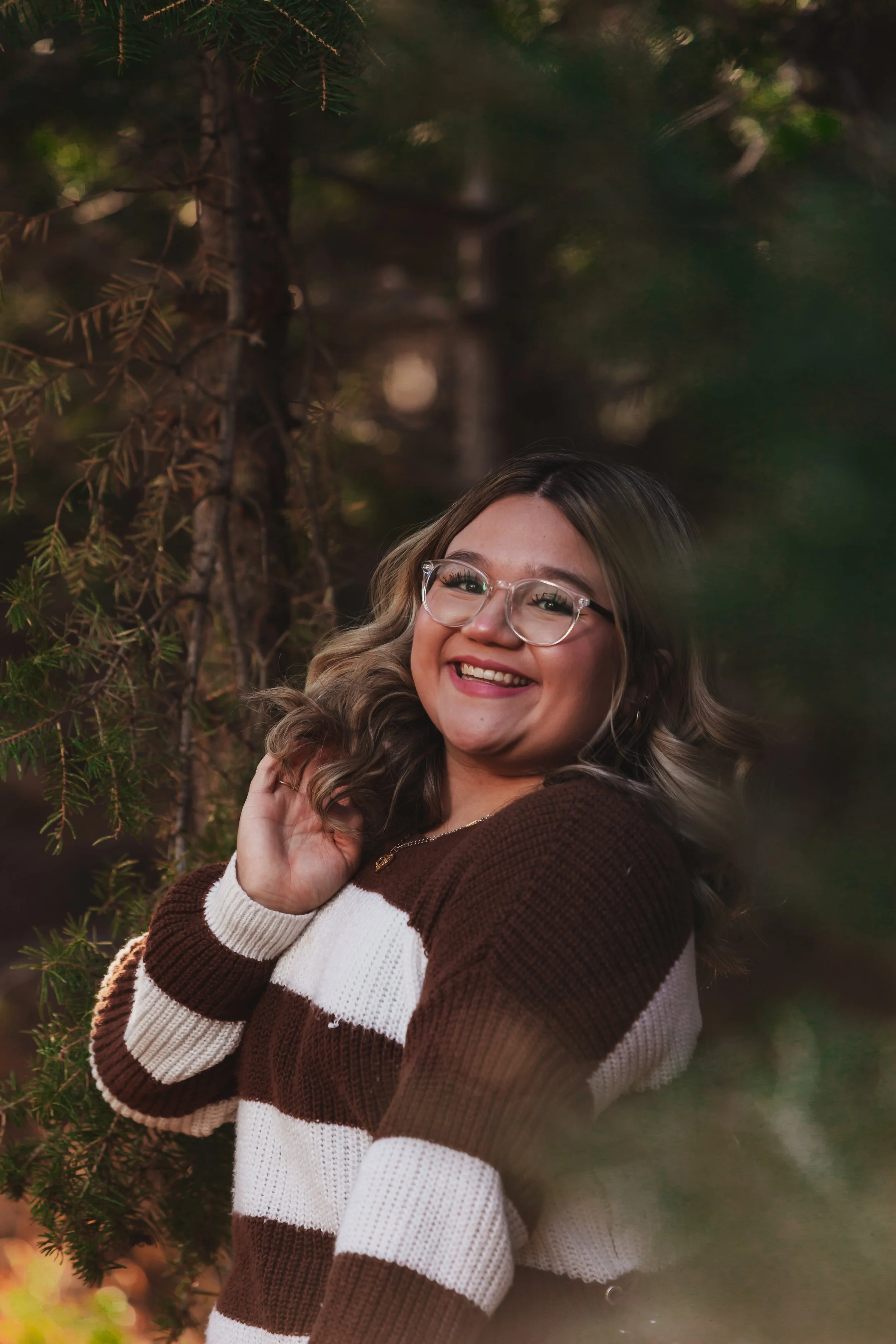 Smiling young woman with wavy hair and glasses in a forest setting, wearing a striped brown and white sweater. Senior Rep photoshoot in Cloudcroft, NM.