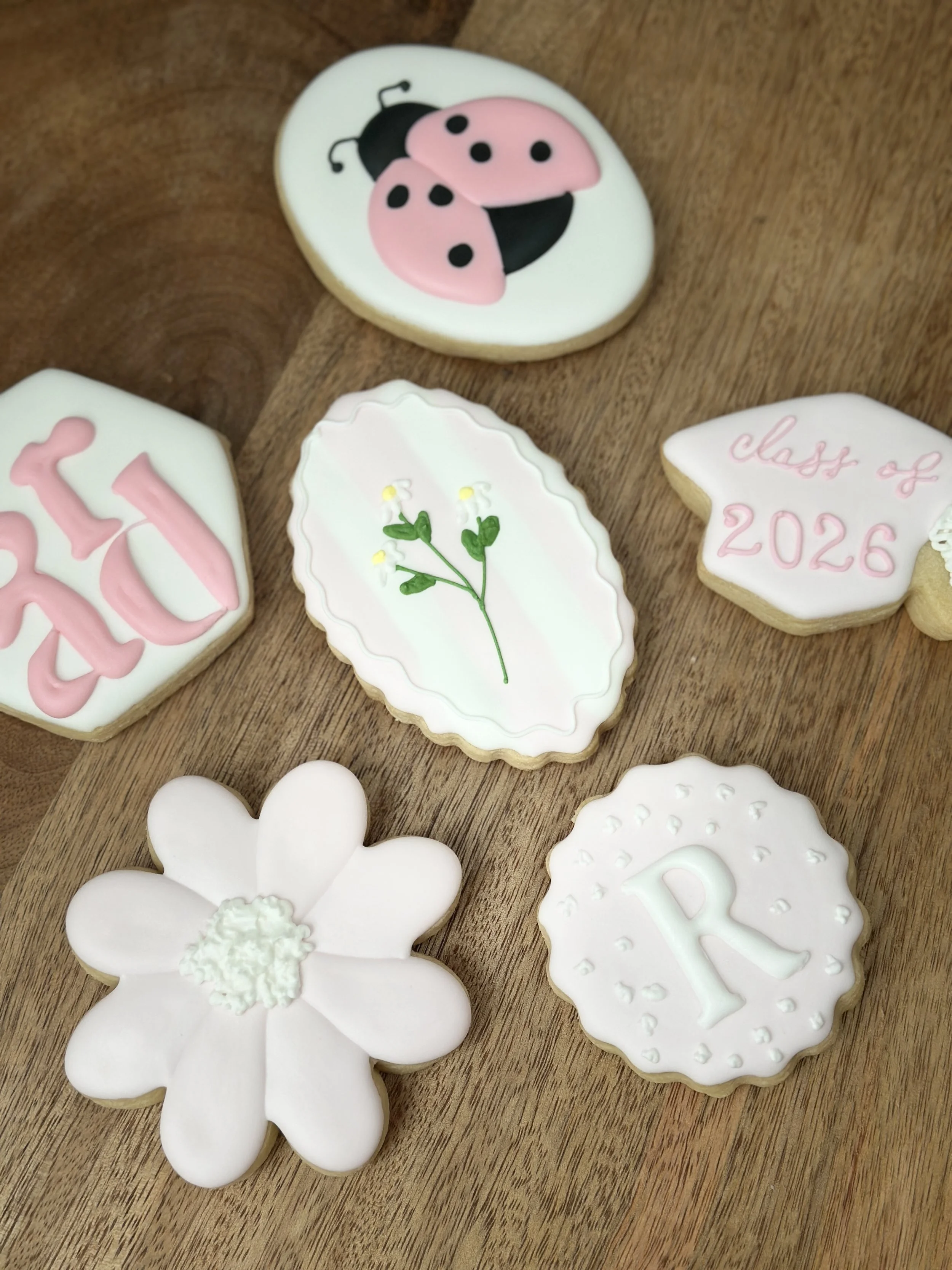 Decorated sugar cookies with various designs on a wooden surface, including a ladybug, a flower, a branch with leaves, and phrases like "class of 2026."