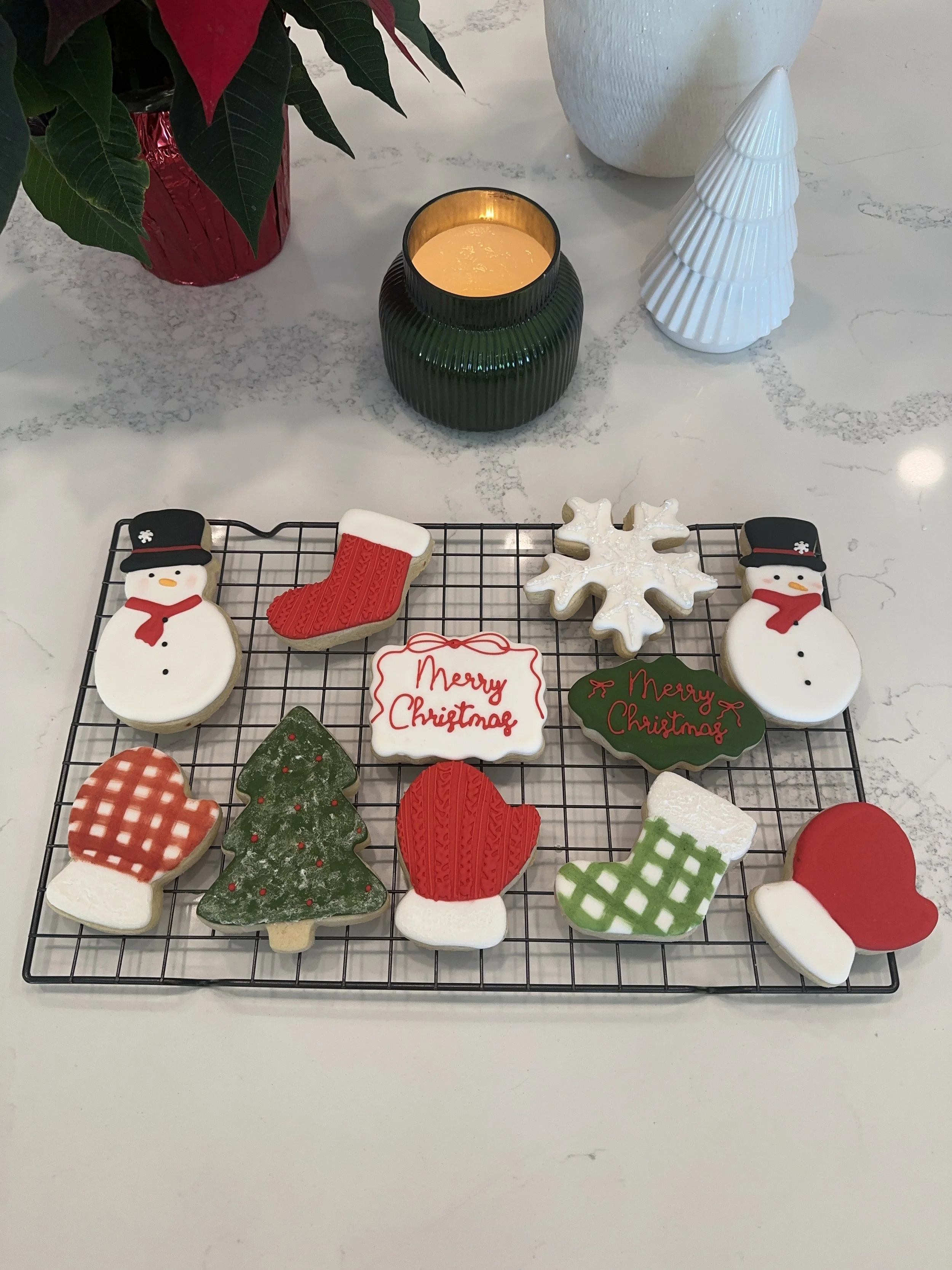 Christmas cookies on a wire rack, including snowmen, Christmas trees, mittens, stockings, and snowflakes, with a "Merry Christmas" message, surrounded by holiday decorations on a white marble surface.