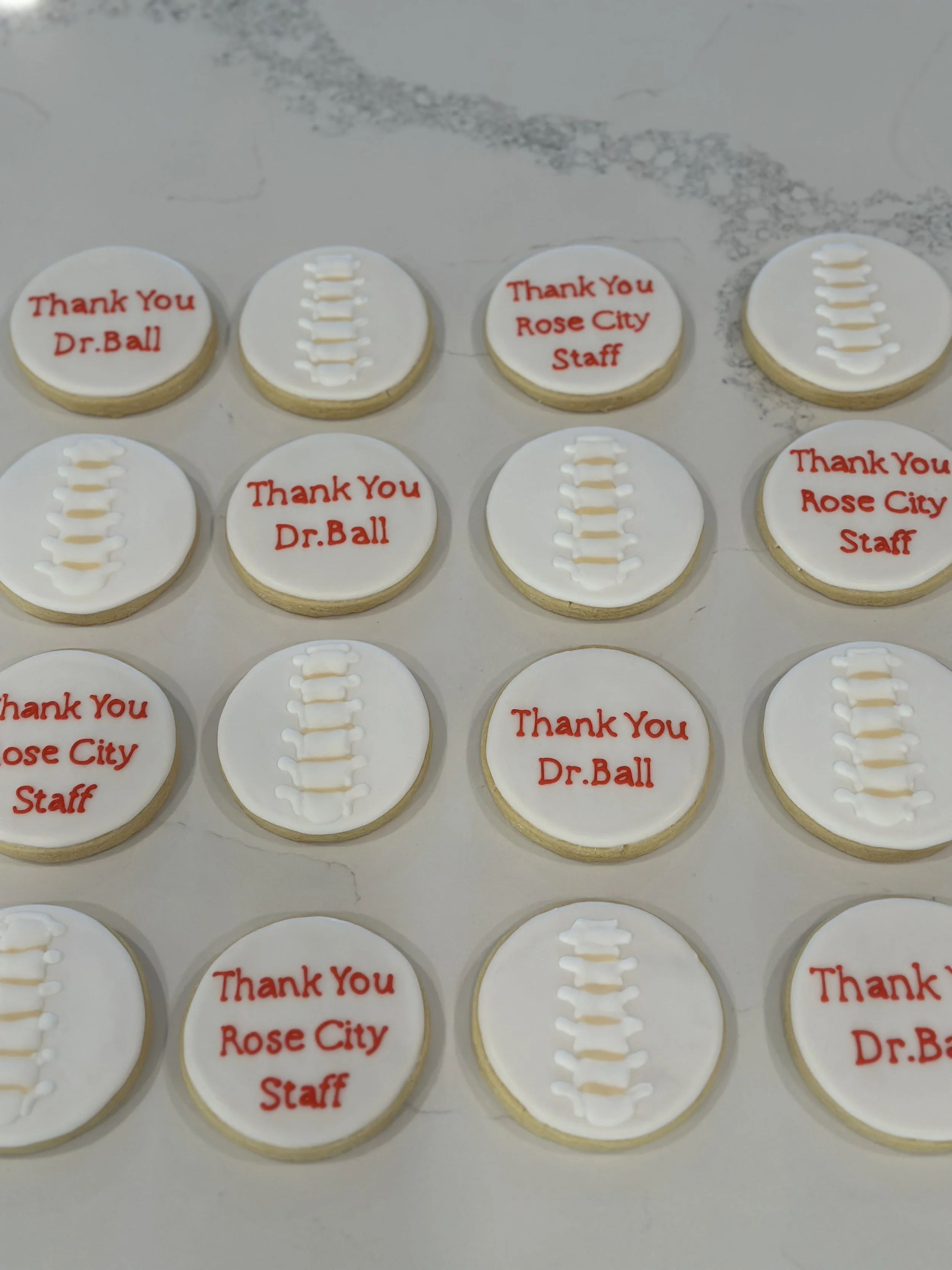 Decorative cookies with white icing, red text saying "Thank You Dr. Ball" and "Thank You Rose City Staff," and a white ladder-like design in the center of some cookies, arranged in rows on a marble surface.