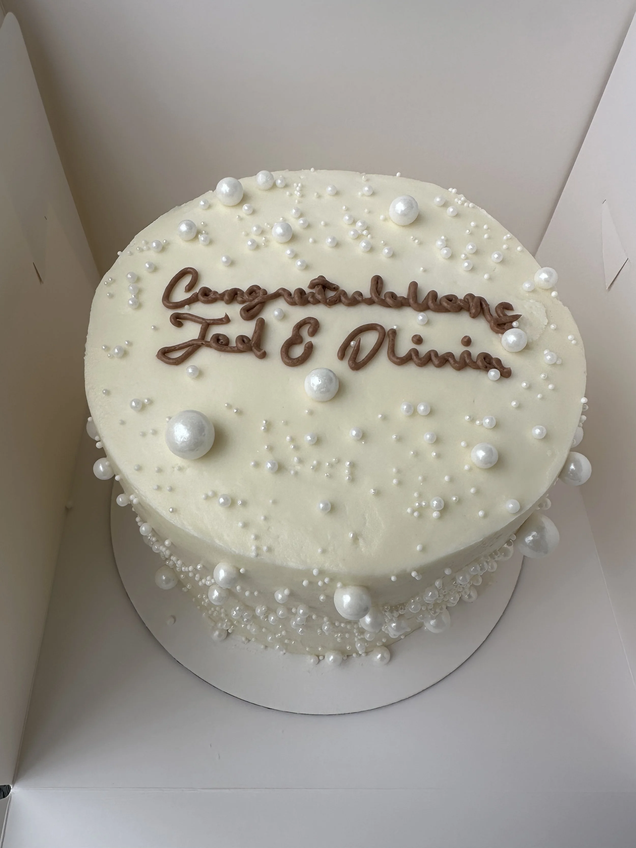 Round white cake decorated with white pearl-like beads and a message written in chocolate that reads 'Congratulations To E. Dumas'. The cake is inside a white box.