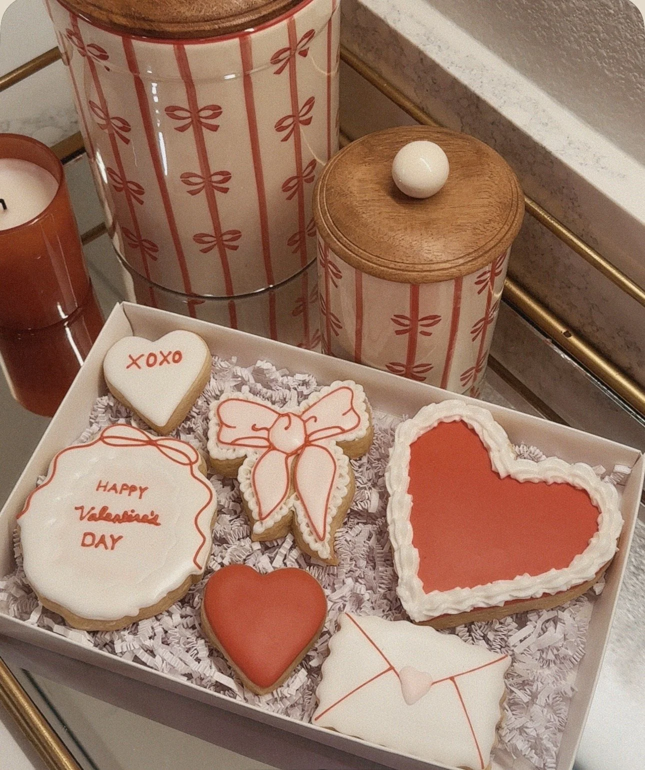 Valentine's Day cookies with red and white icing, including hearts, a bow, and one with the message 'Happy Valentine's Day' in a gift box, with candles and decorative jars in the background.