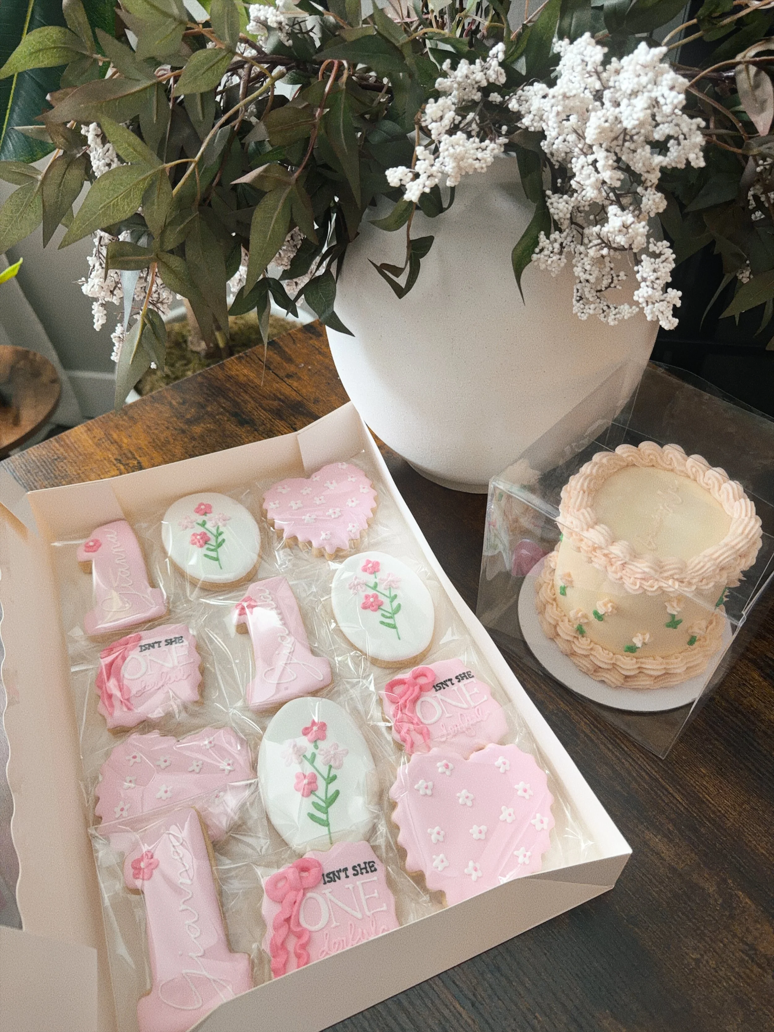 A white box filled with pink and white decorated cookies in various shapes, including hearts and flowers, with some cookies inscribed with messages like 'You Are Loved' and 'You Are Enough'. Beside the box is a large white vase with greenery and whit