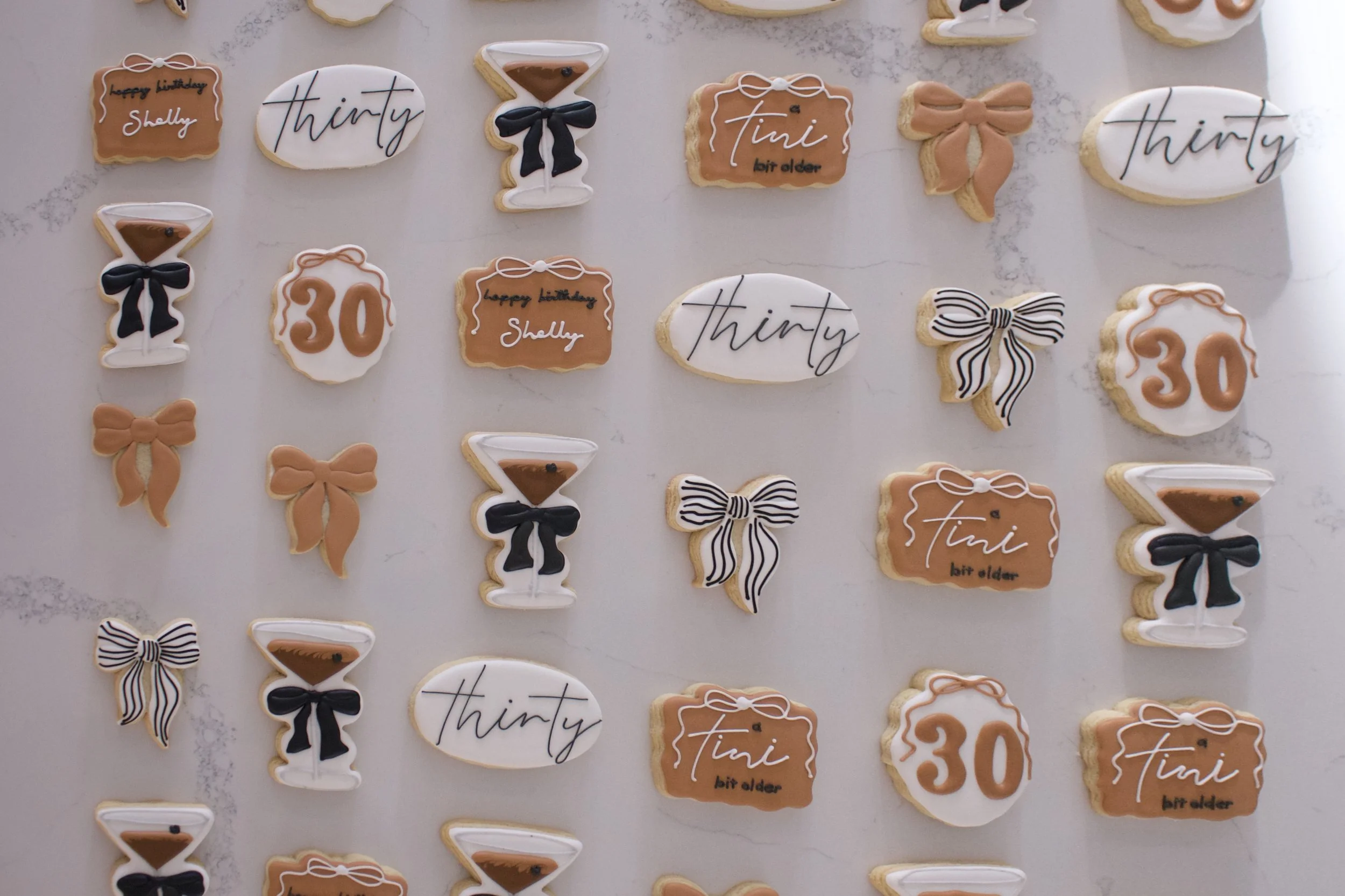 Collection of decorated birthday cookies including ones with the words 'happy birthday Shelly,' 'thirty,' and '30,' as well as cookies shaped like bows, cocktail glasses, and the word 'tini,' on a white surface.