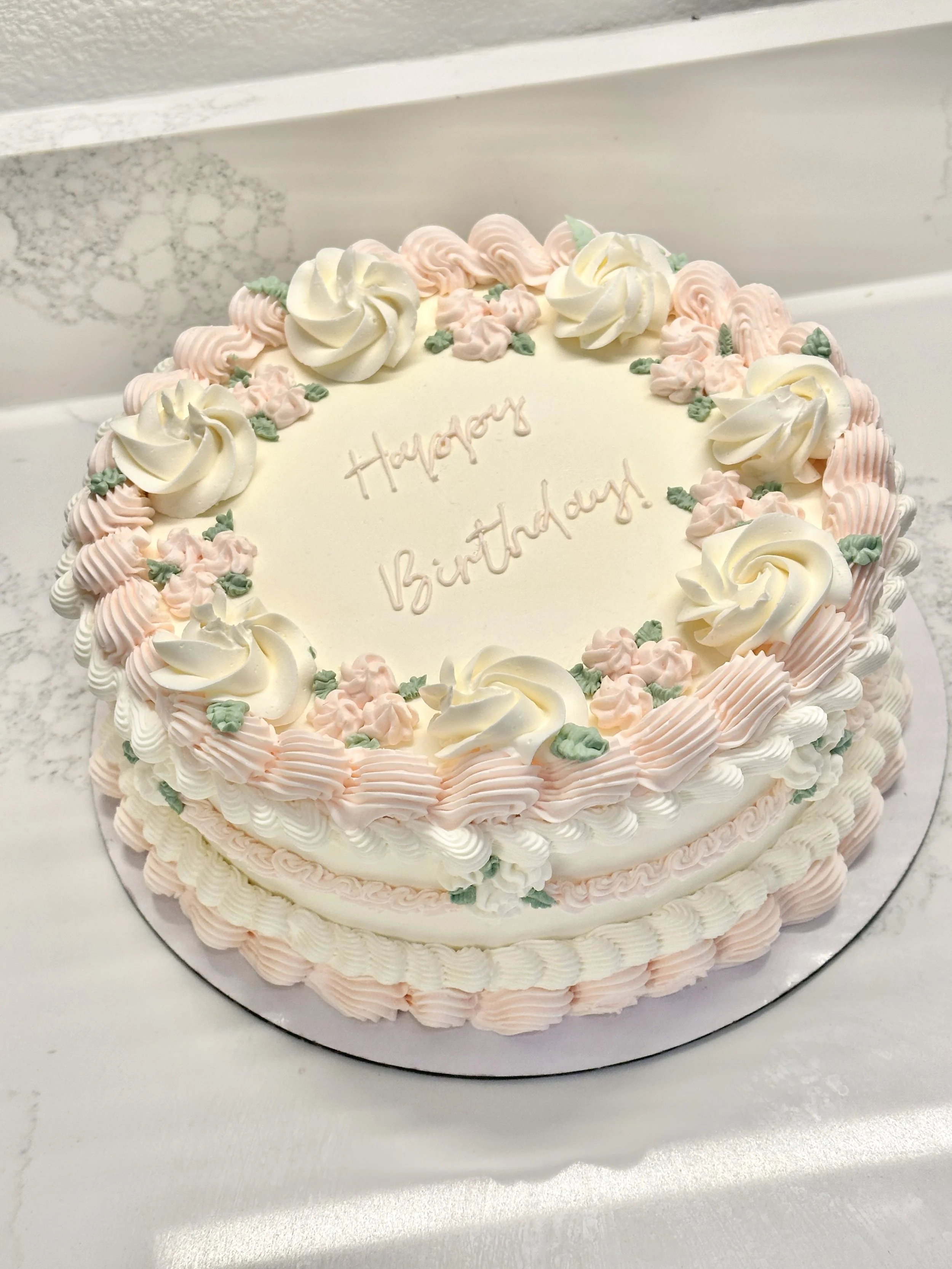 A round birthday cake decorated with pink and white icing flowers and swirls, with the words 'Happy Birthday!' written on the top in light pink icing.