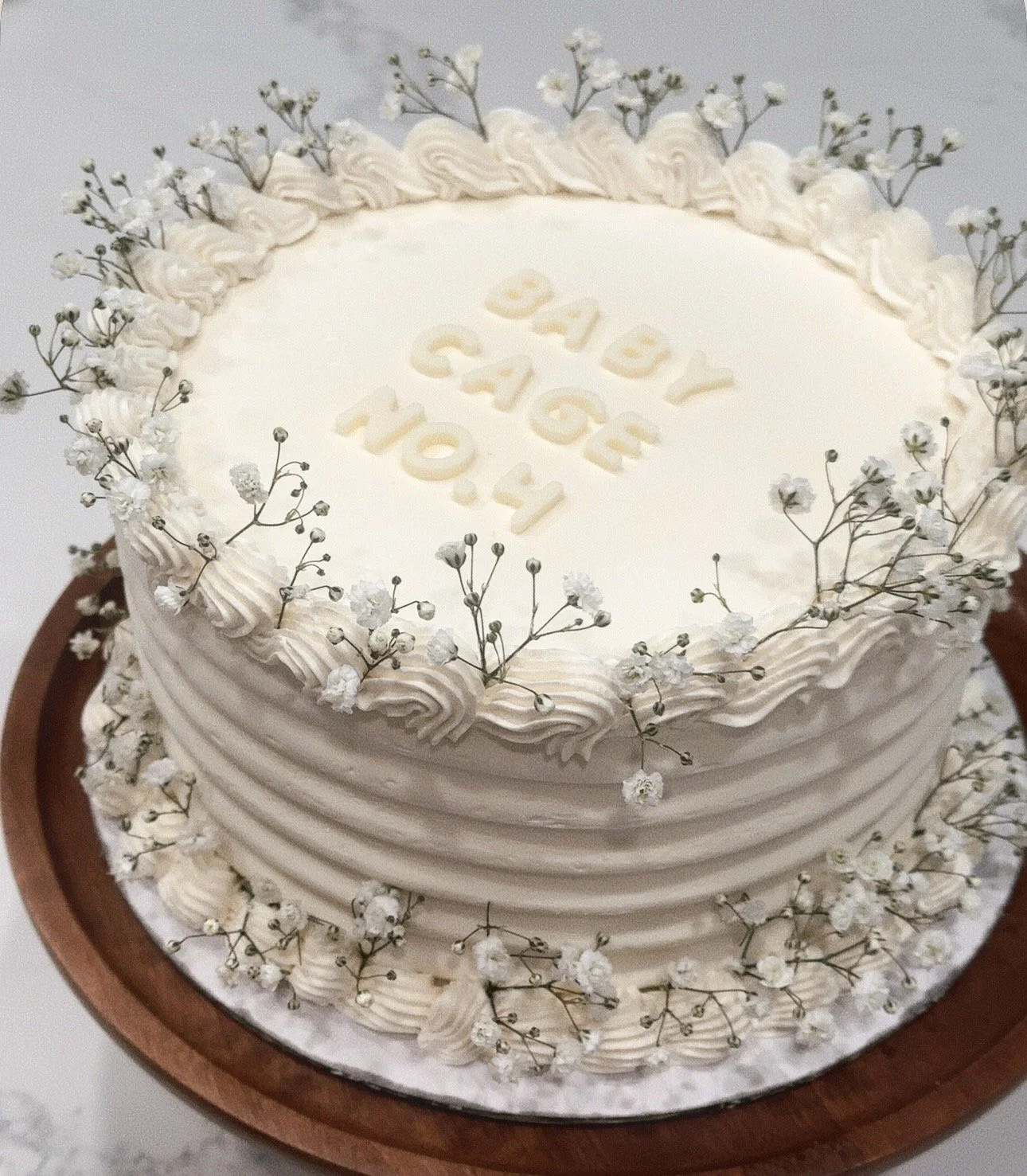 A white birthday cake with piped frosting decorations, small white flowers, and the words 'Happy Birthday' on top, placed on a round wooden cake stand.