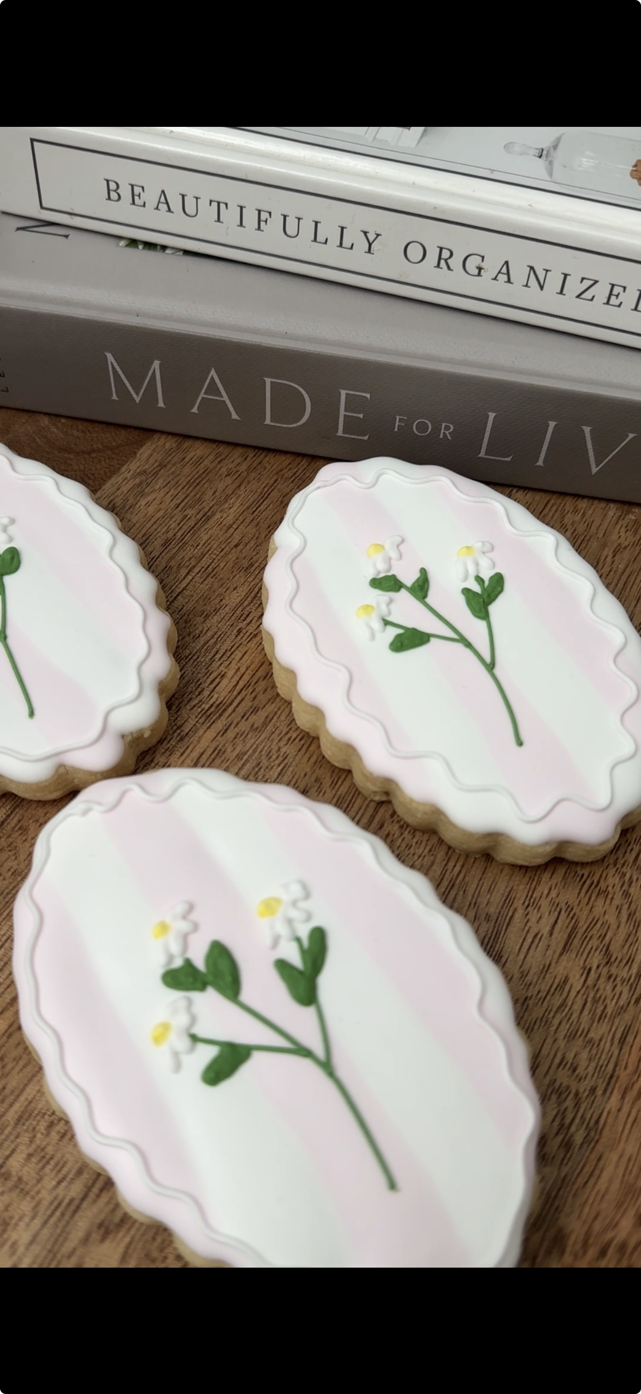 Decorated cookies with white icing and a floral design, placed on a wooden surface with books in the background.