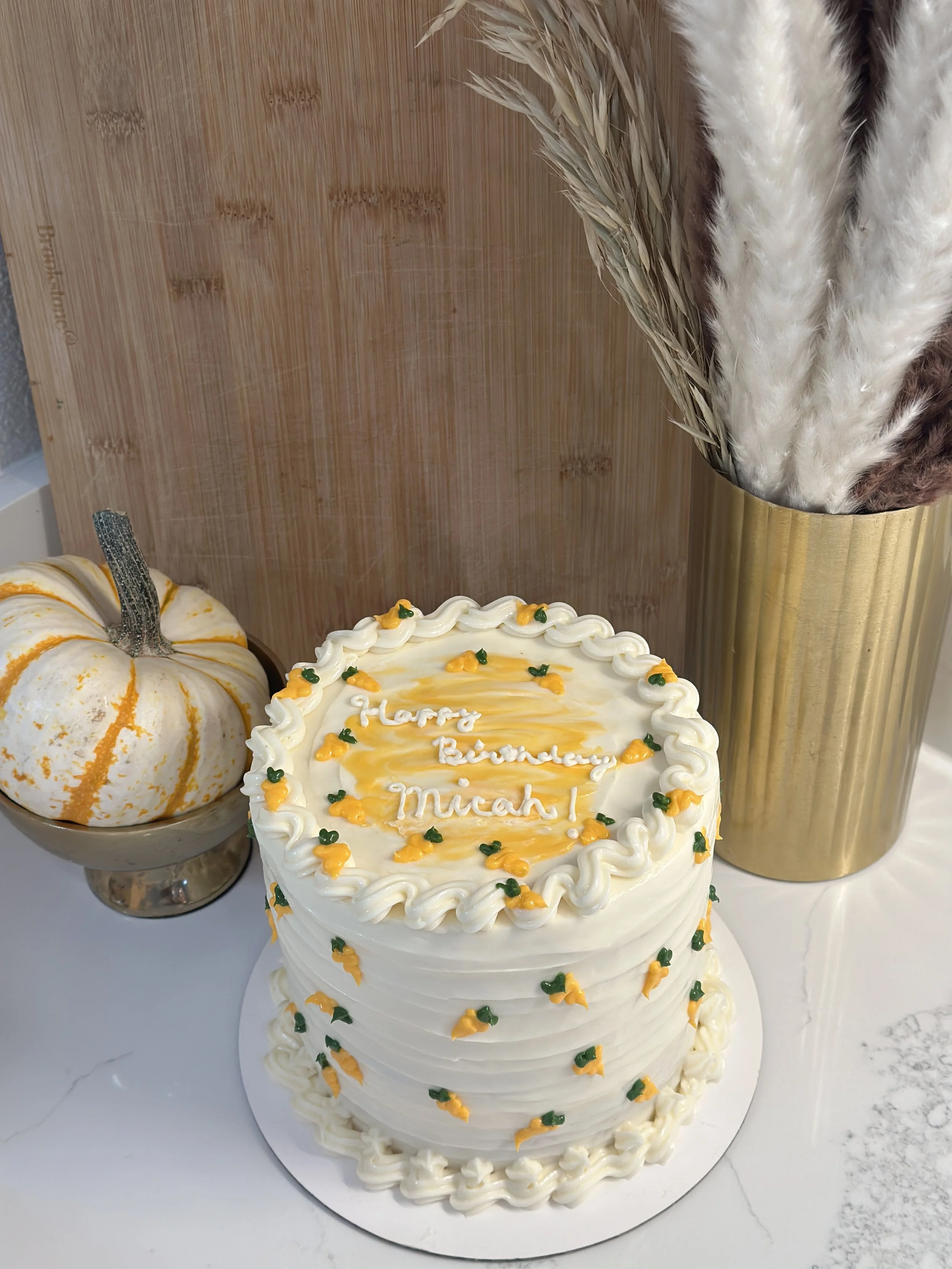 A birthday cake with white frosting, decorated with small yellow and green embellishments, and the words 'Happy Birthday Micah' written on top, alongside a small pumpkin-shaped cake with white and orange icing, and a tall vase holding decorative drie