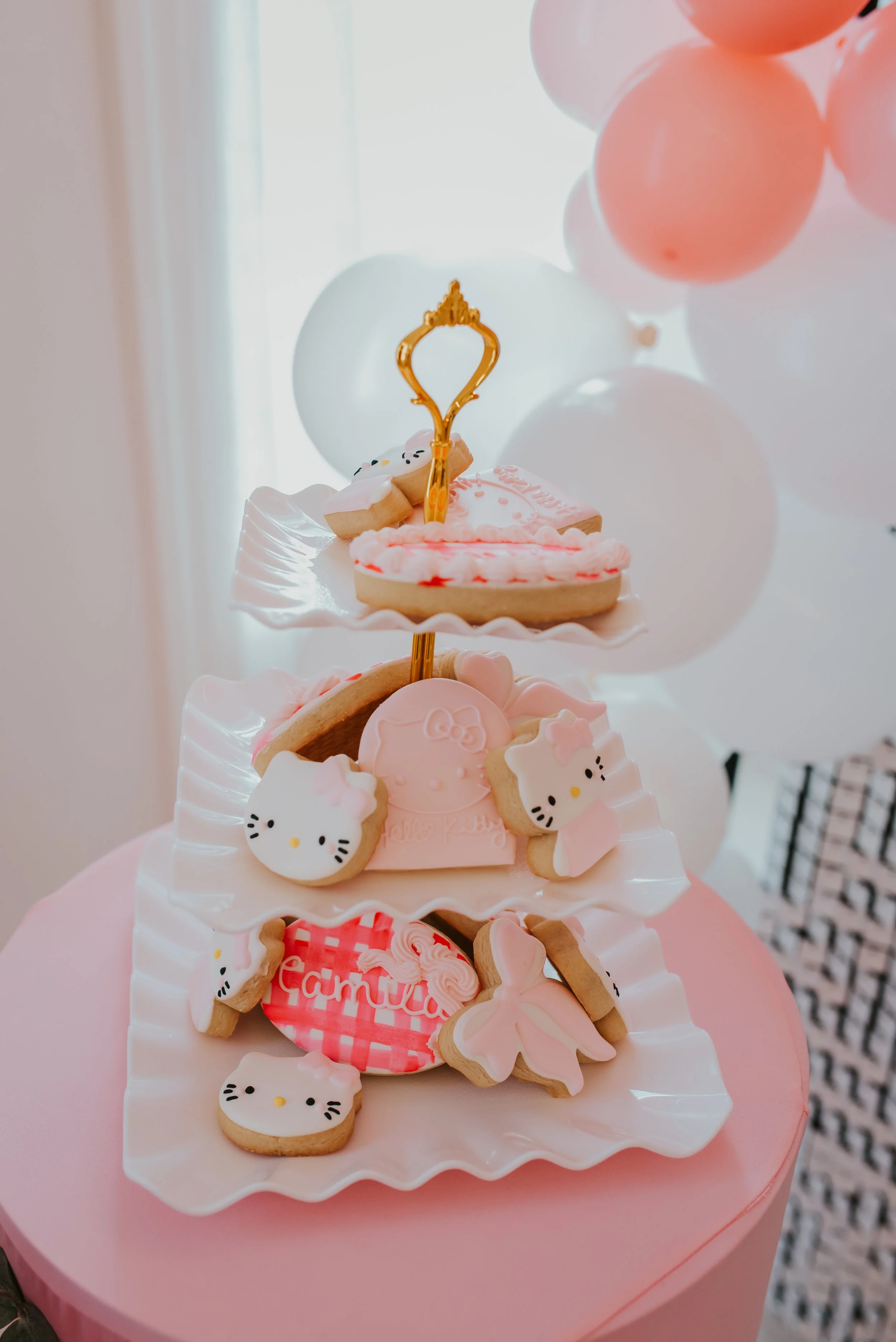 A tiered pink and white cake decorated with cookies shaped like Hello Kitty, leaves, and pink themes, with balloons in the background.