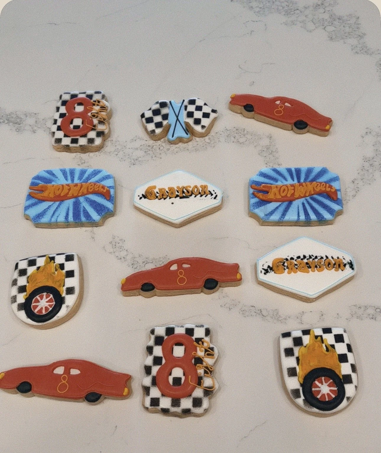A collection of decorated cookies with racing and car themes, including checkered flags, cars, race-related words like "Hot Wheels" and "Crayson," and racing numbers.