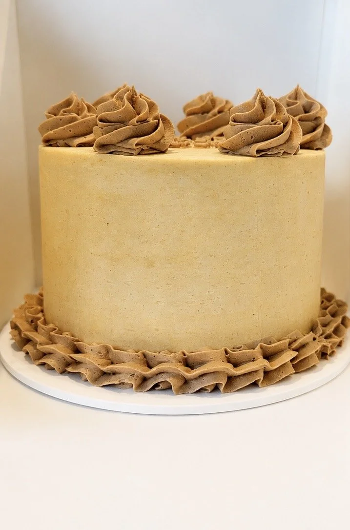 A tall, round cake decorated with beige frosting, featuring rosette swirls on top and piped border at the base.