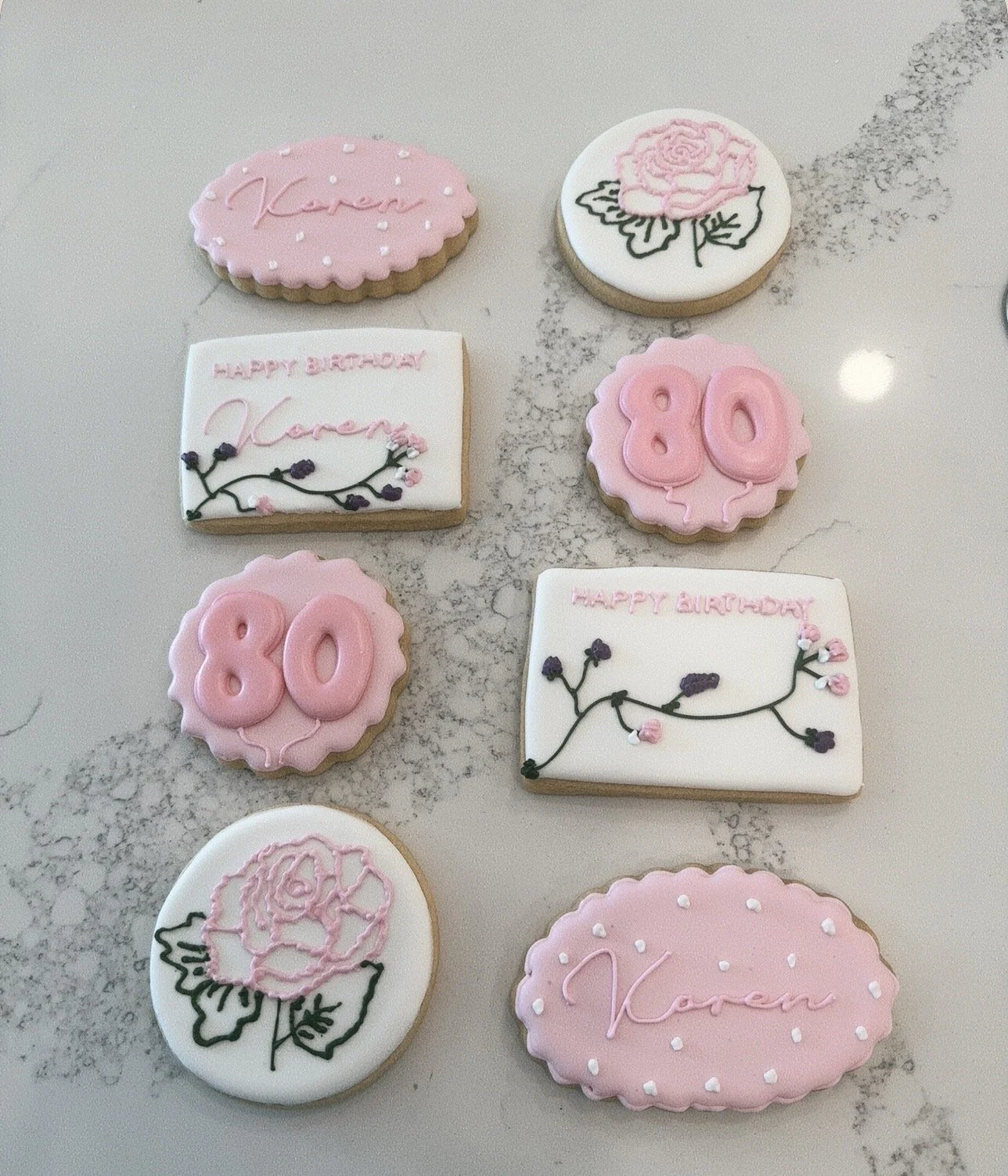 Decorated sugar cookies with pink, white, and black icing, featuring the words 'Karen,' 'happy birthday,' and the number '80,' along with floral designs of roses and branches.