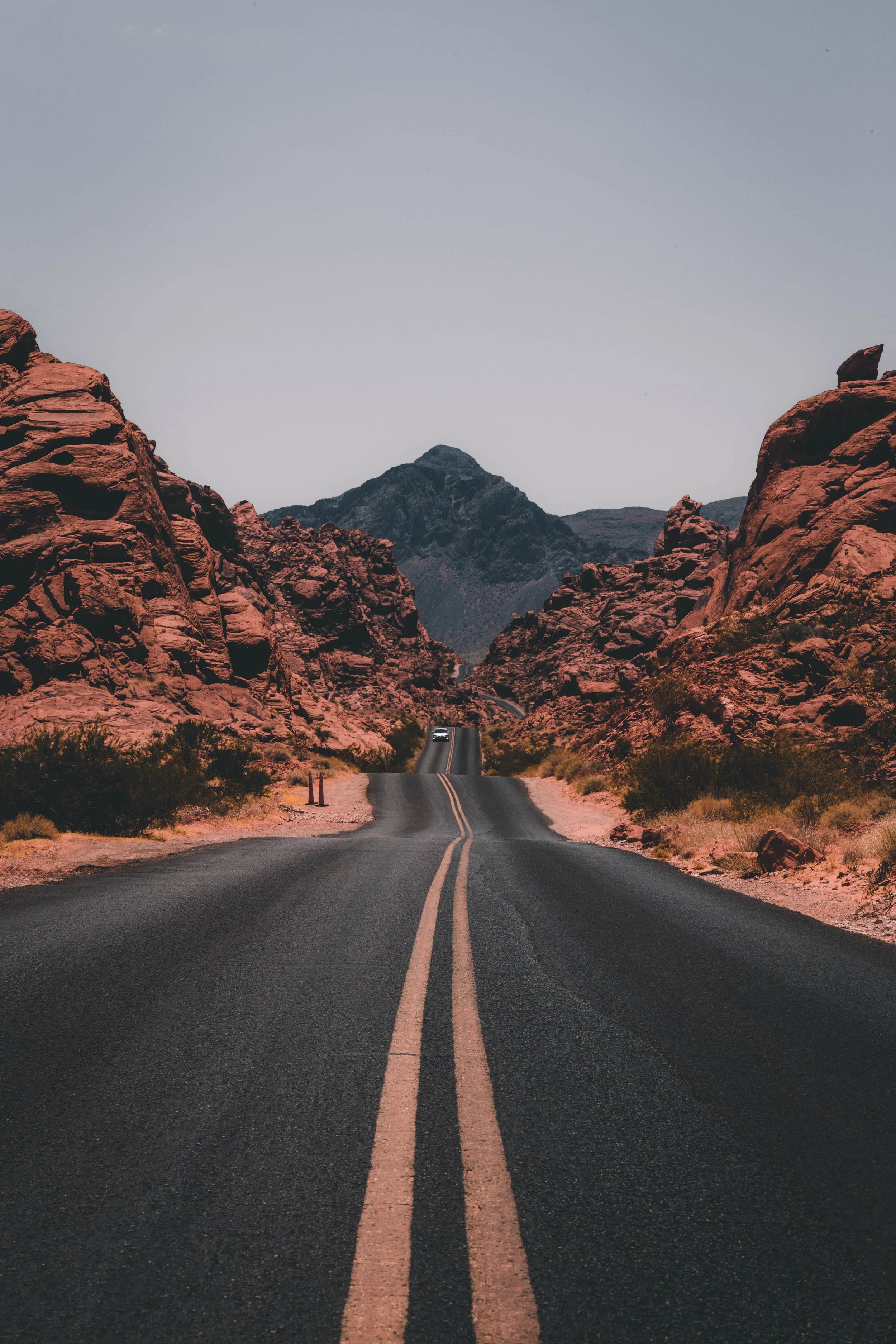 A straight black asphalt road with a yellow double line running through a desert landscape with red rocks and mountains in the background under a clear sky.
