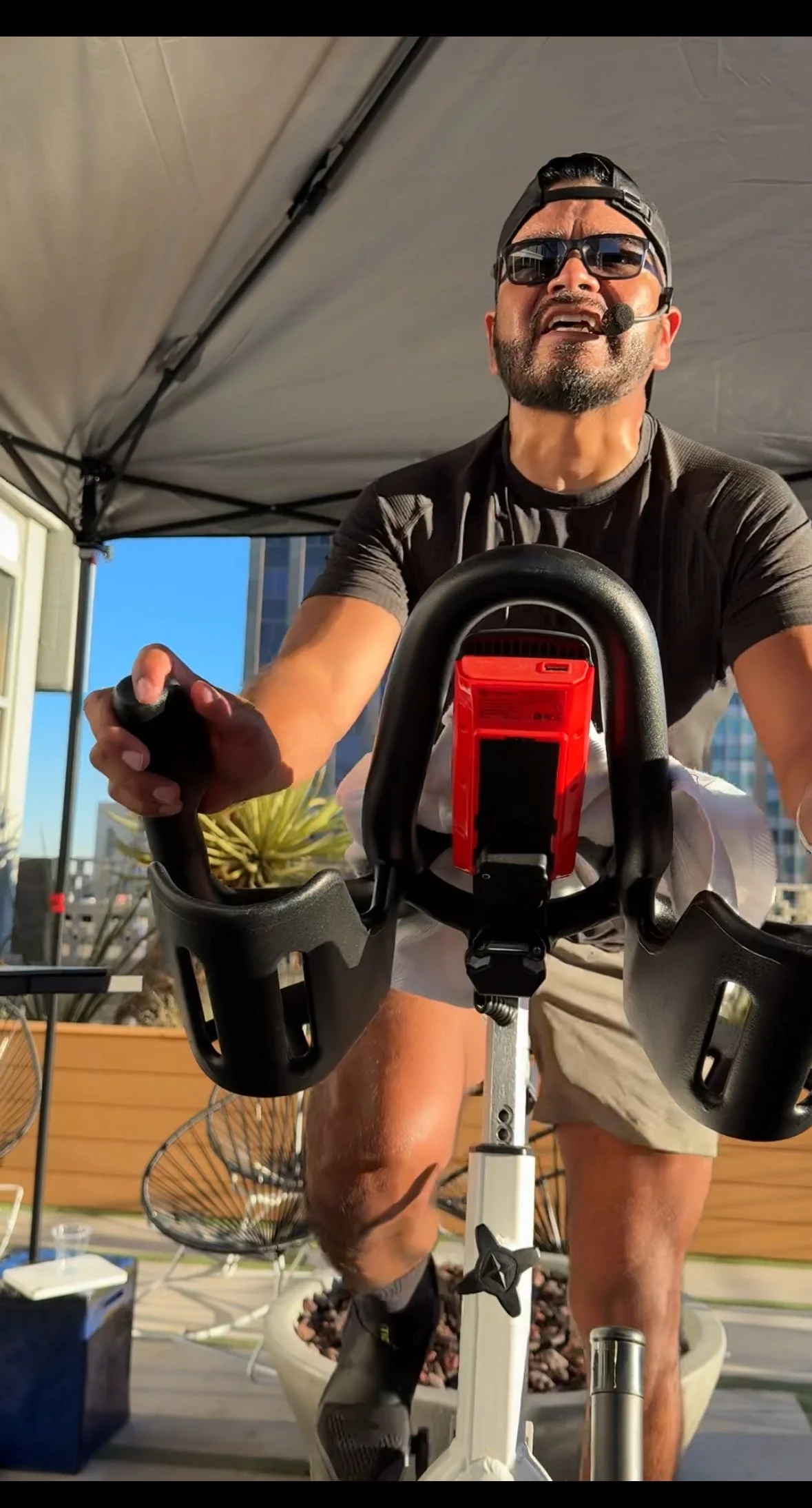 Man wearing sunglasses and a cap riding a stationary exercise bike indoors, speaking into a headset microphone, with plants and chairs in the background.