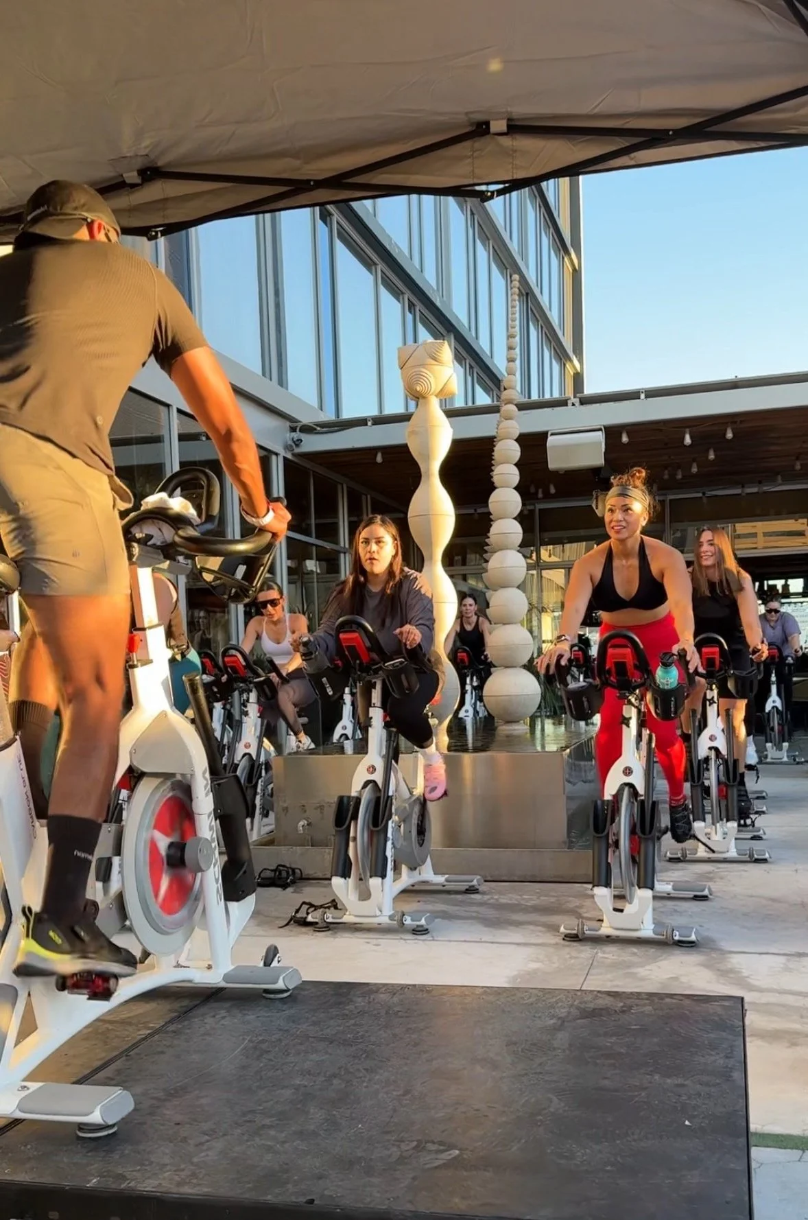 Outdoor spin class with instructor and participants on stationary bikes under canopy.