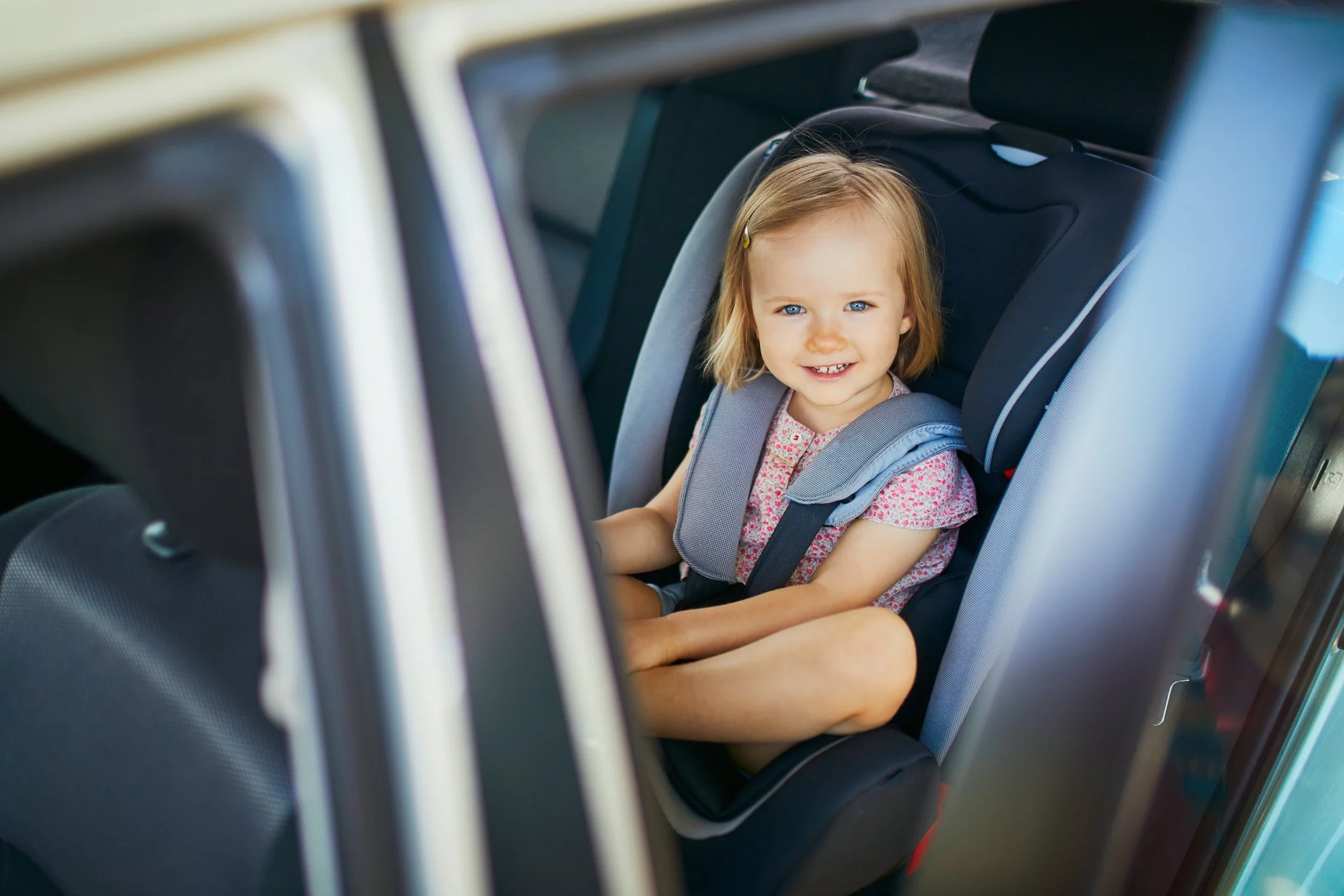 A young girl with blonde hair and blue eyes sitting in a child's car seat inside a vehicle, smiling at the camera.