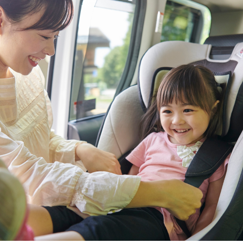 A smiling young girl sits in a car seat while a woman, possibly her mother, helps secure her in a seat belt inside a vehicle.
