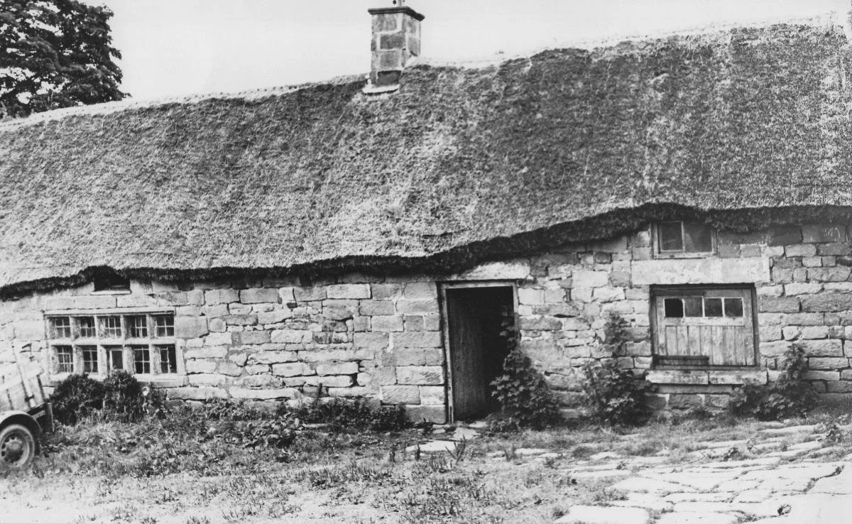 Black and white photo of an old stone cottage with a thatched roof, small windows, a central door, and some plants growing outside.