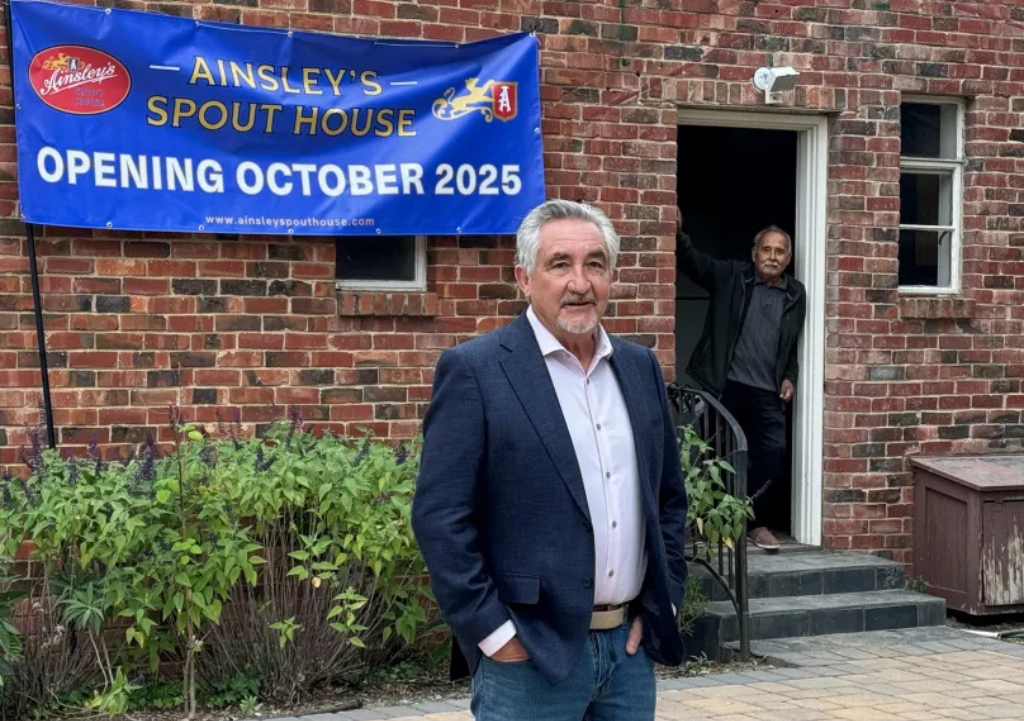 Two men standing outside a brick building with a blue banner that reads "Ainsley's Spout House Opening October 2025." One man in a suit coating stands in the foreground, while the other man stands in the doorway in the background.