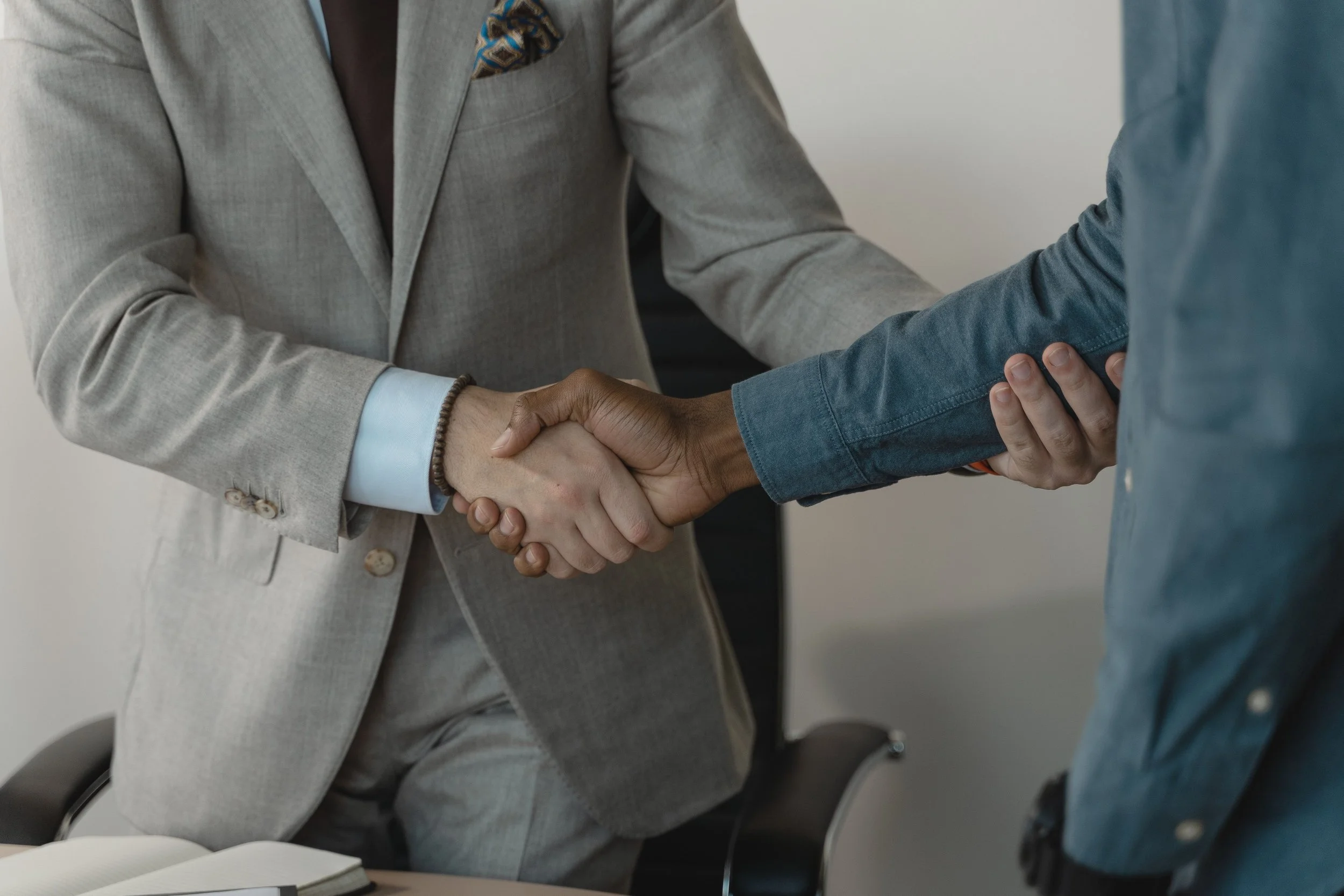 A close-up of two people shaking hands during a business meeting, with one person wearing a beige suit and the other wearing a blue shirt.