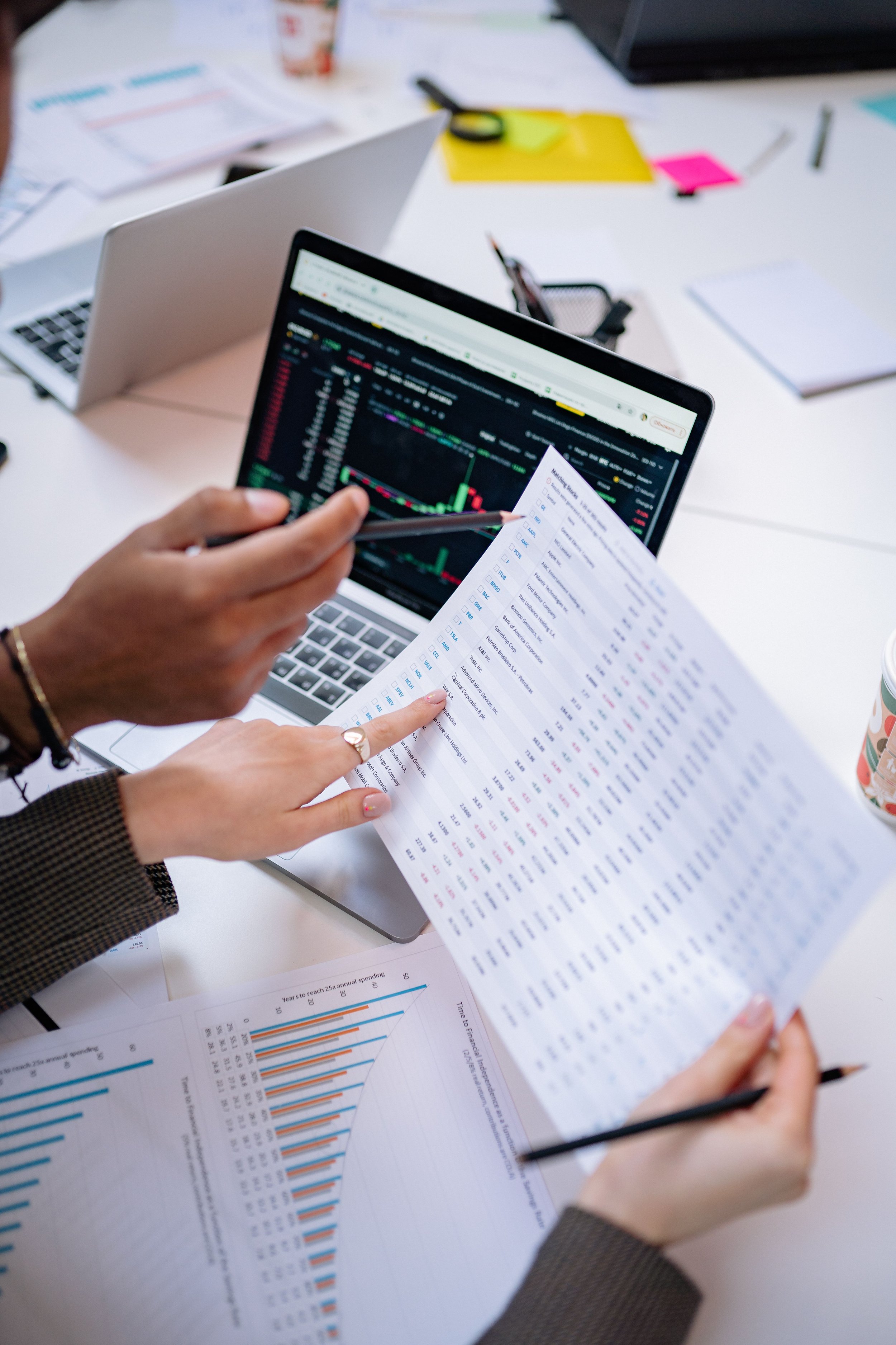 Work desk with multiple papers, charts, and laptops, with people analyzing financial data and market graphs.