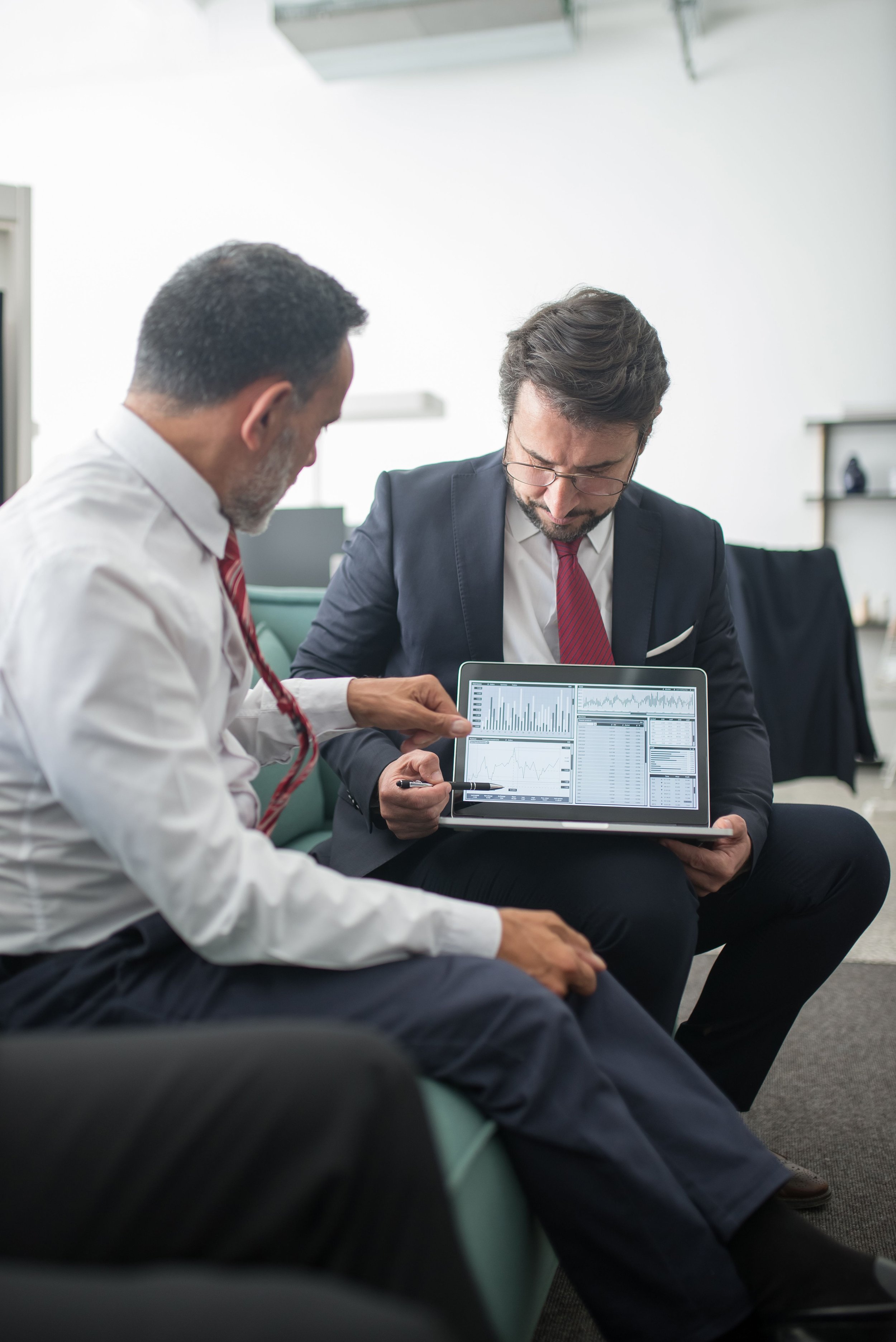 Two men in business attire sitting in a waiting area, looking at a tablet displaying graphs and charts.