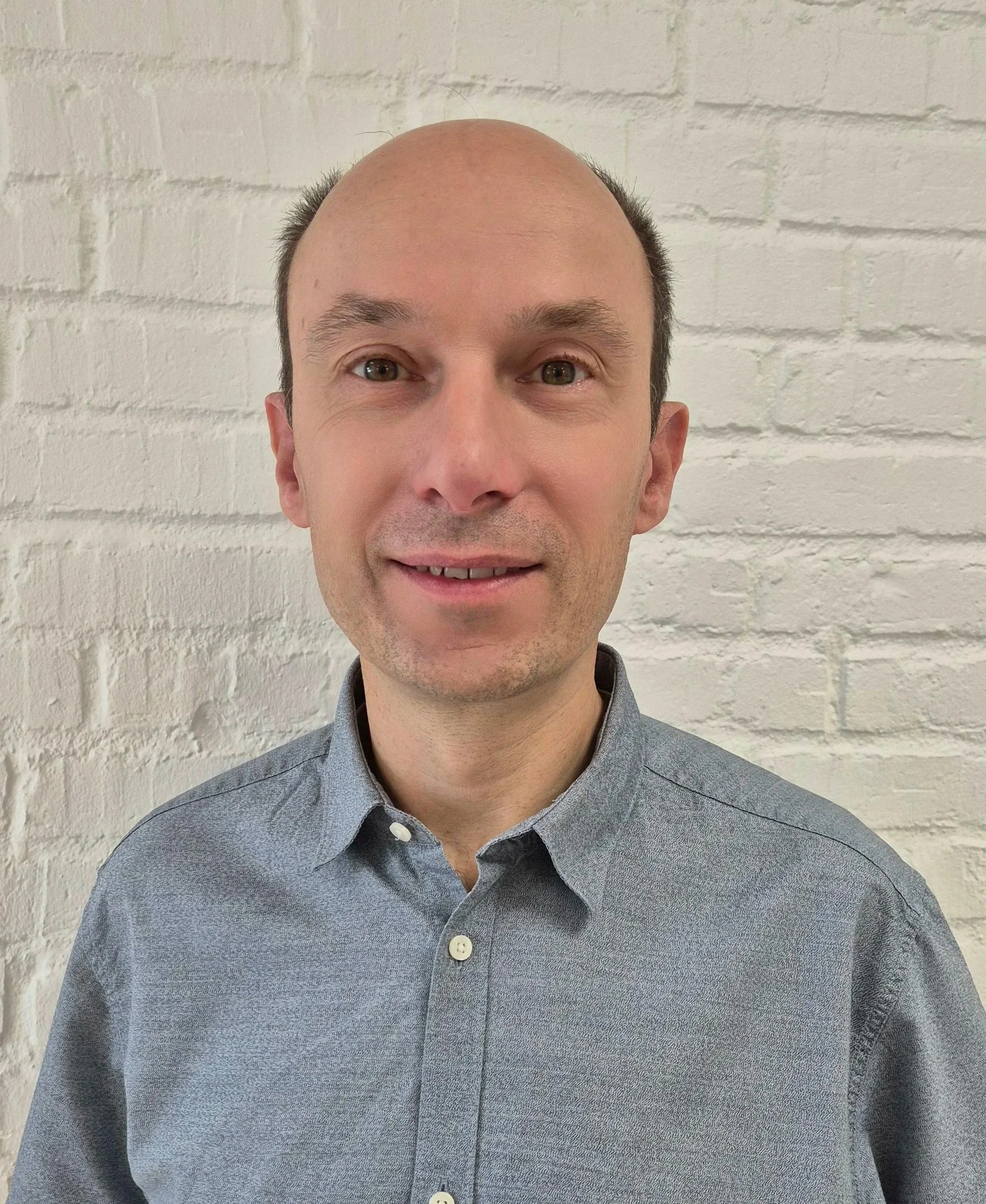 A man with a balding head, wearing a gray button-up shirt, standing against a white brick wall.