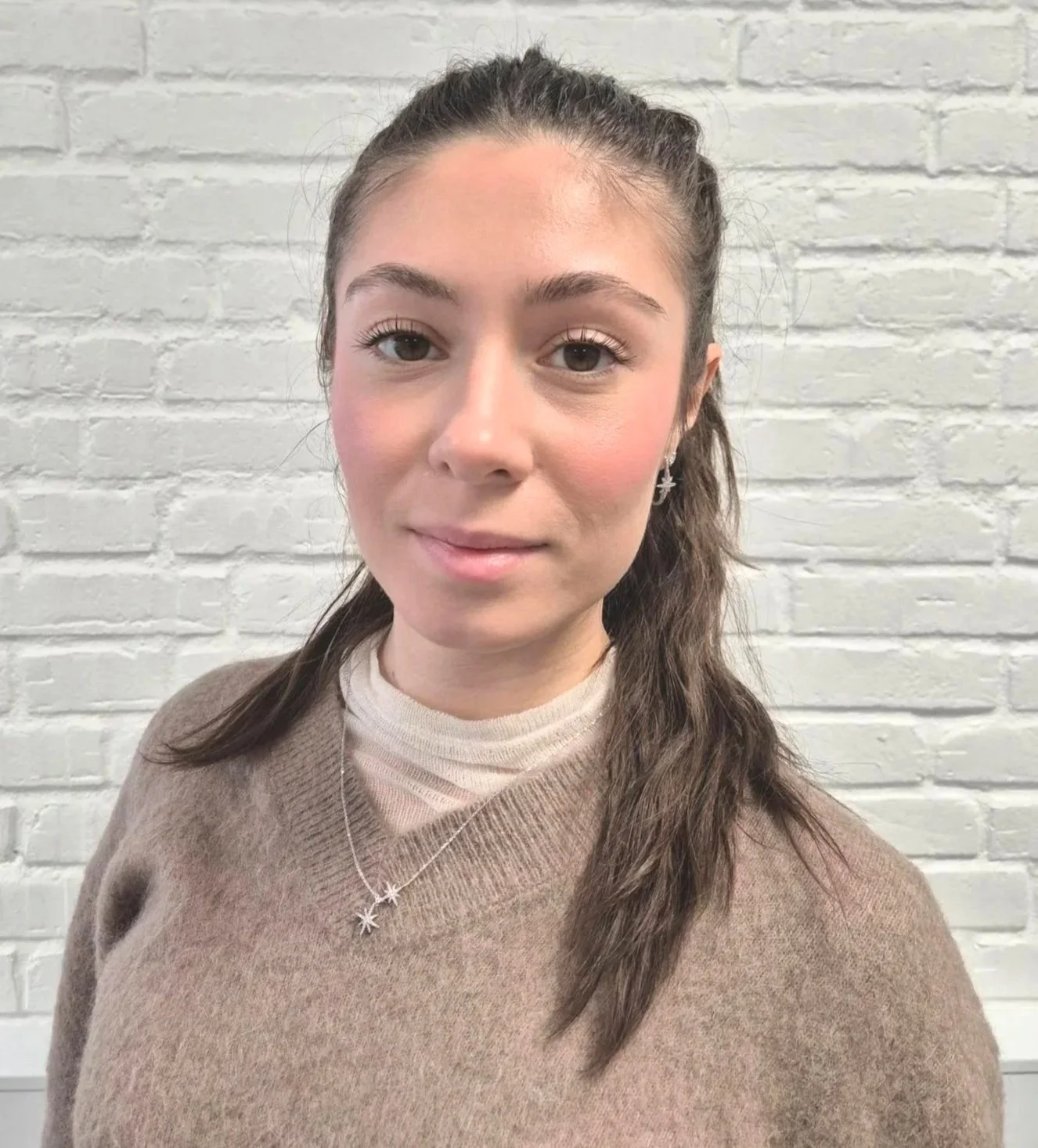 A young woman with light brown hair in a braid, wearing a beige sweater over a white top, standing in front of a white brick wall.