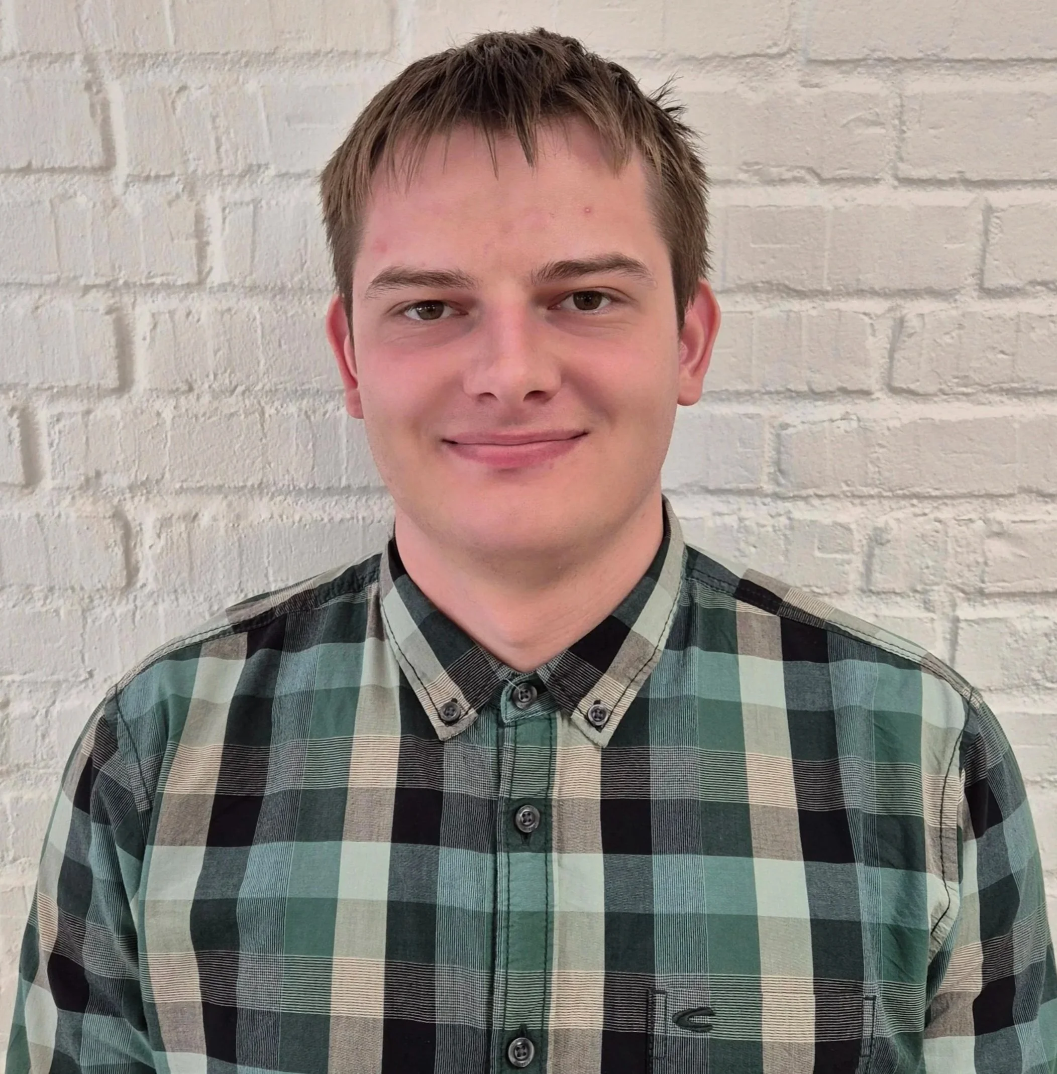 A young man with short brown hair, fair skin, and a slight smile standing in front of a white brick wall, wearing a green and black plaid button-up shirt.