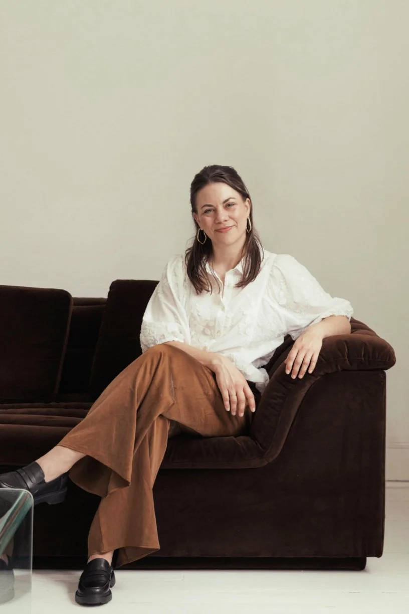 A woman with dark brown hair wearing a white blouse, brown pants, and black shoes sitting on a dark brown velvet sofa in a room. Hobart based copywriter for creative founders.