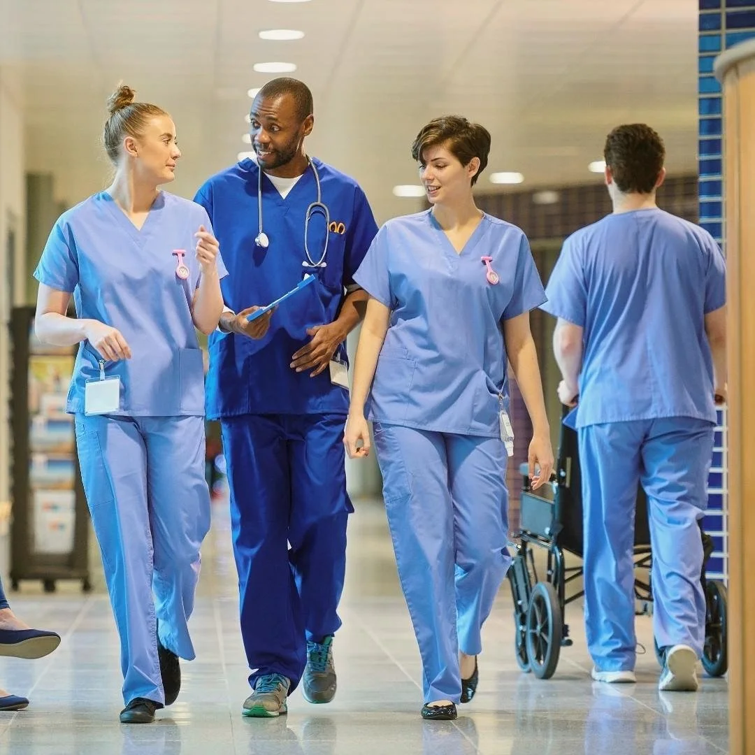 Pflegekräfte und ein Arzt im Gespräch auf dem Flur einer Klinik, im Hintergrund ein Rollstuhl.
