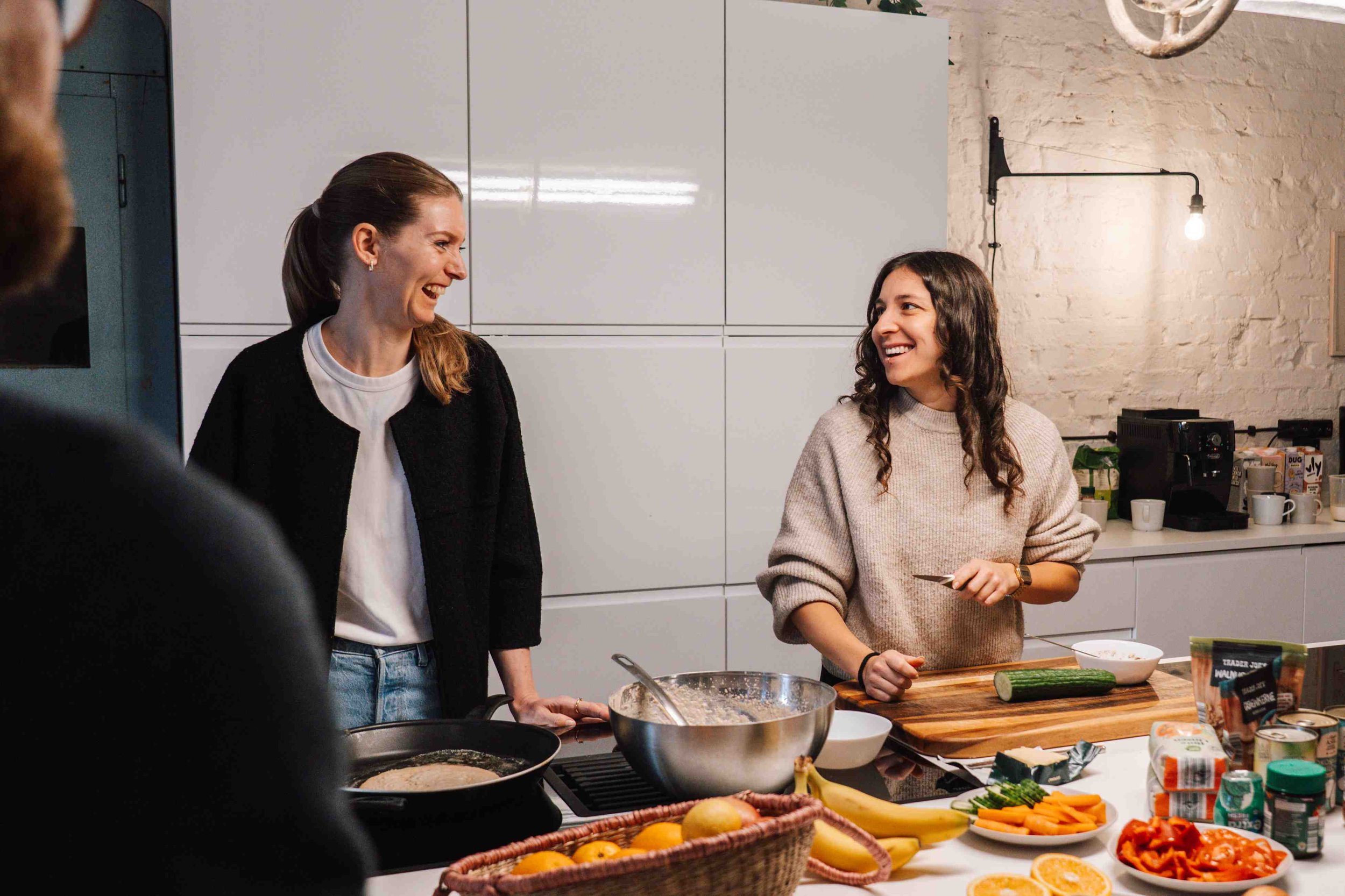 Zwei Frauen kochen gemeinsam lachend in einer Küche mit frischem Obst und Gemüse