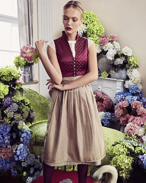 Female Model in dunkelrotem Dirndl mit weißer Bluse und beiger Schürze, posiert vor Blumenvasen voller bunter Hortensien. Kampagnenaufnahme von imagespy für die Trachtenmanufaktur München.