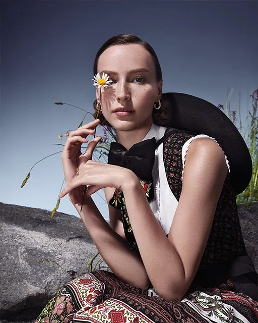 Model in traditioneller Tracht hält eine Margerite vors Gesicht, sitzt auf einem Stein vor einer hohen Wiese – zeitgemäße Trachteninszenierung von imagespy für Vogue und Lodenfrey.