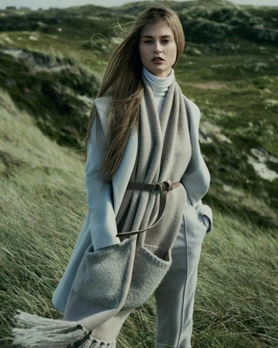 Hessnatur Advertorial Bild eines Models vor Hügellandschaft in hellblauem Mantel, Rollkragenpullover, grauer Hose und Fransenschal mit Hand in der Schaltasche; umgesetzt von imagespy